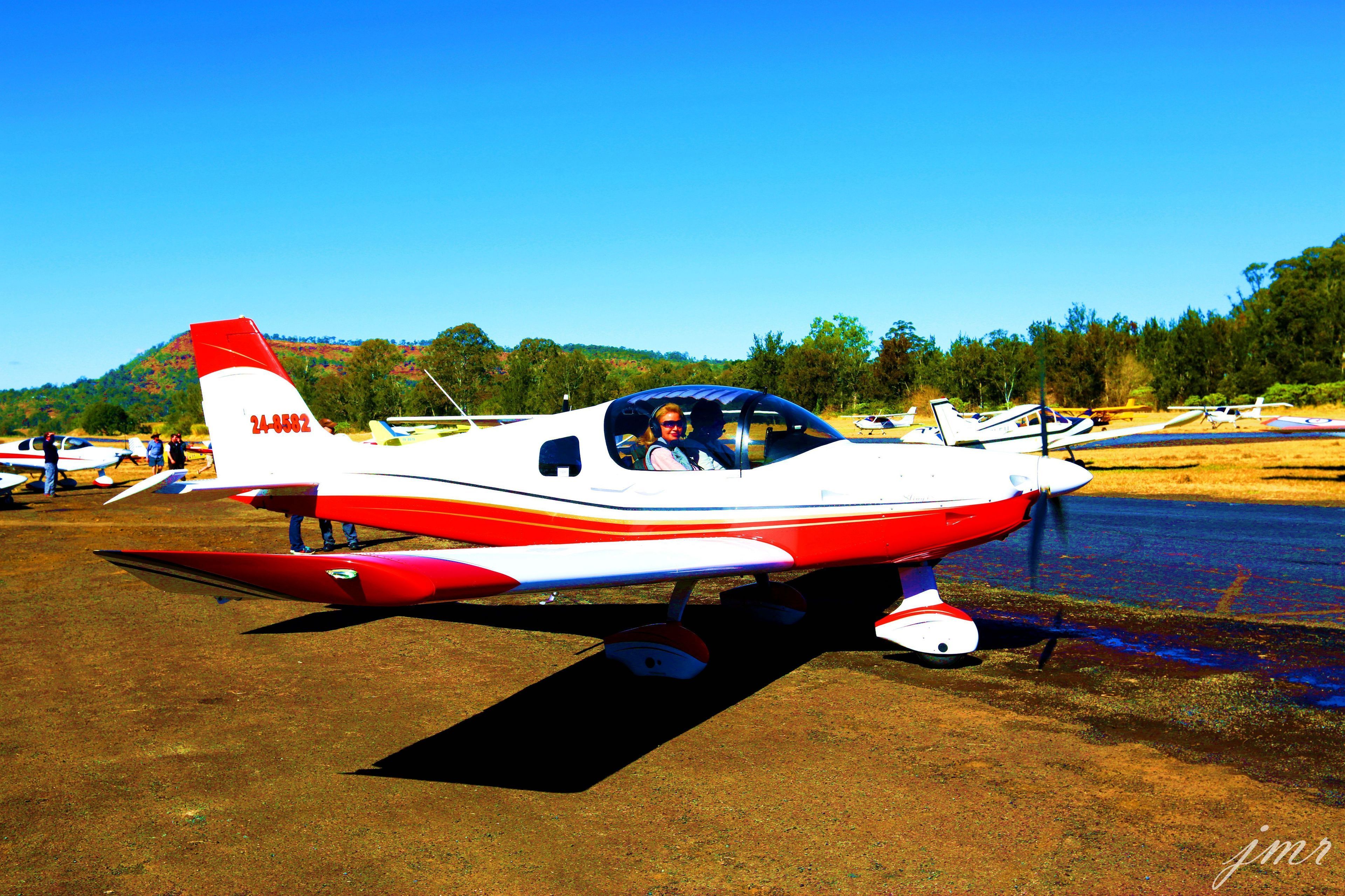 Un petit avion rouge et blanc est garé sur une surface de terre, avec un ciel bleu dégagé et des arbres en arrière-plan.