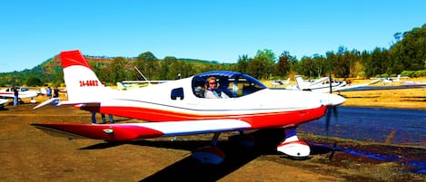 Un petit avion rouge et blanc est garé sur une surface de terre, avec un ciel bleu dégagé et des arbres en arrière-plan.