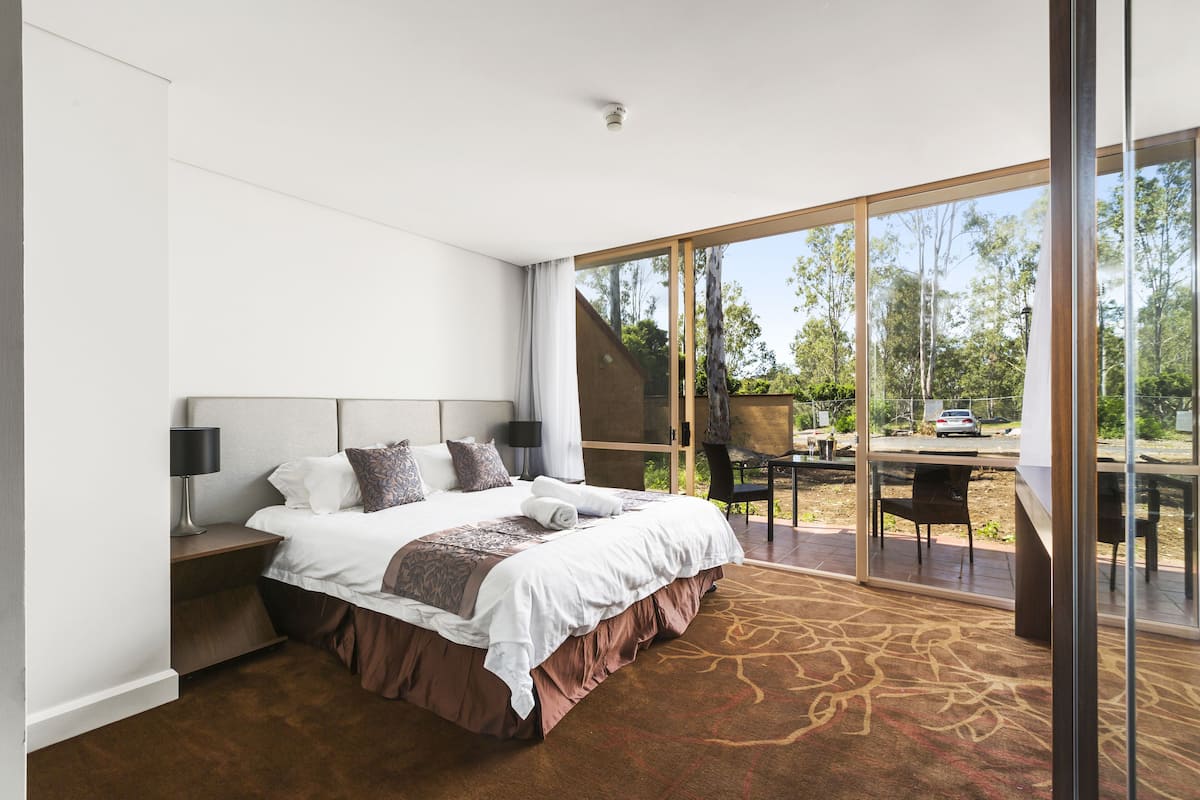 A modern hotel room with a large bed, a nightstand, a desk, and a view of the outdoors through large glass doors.