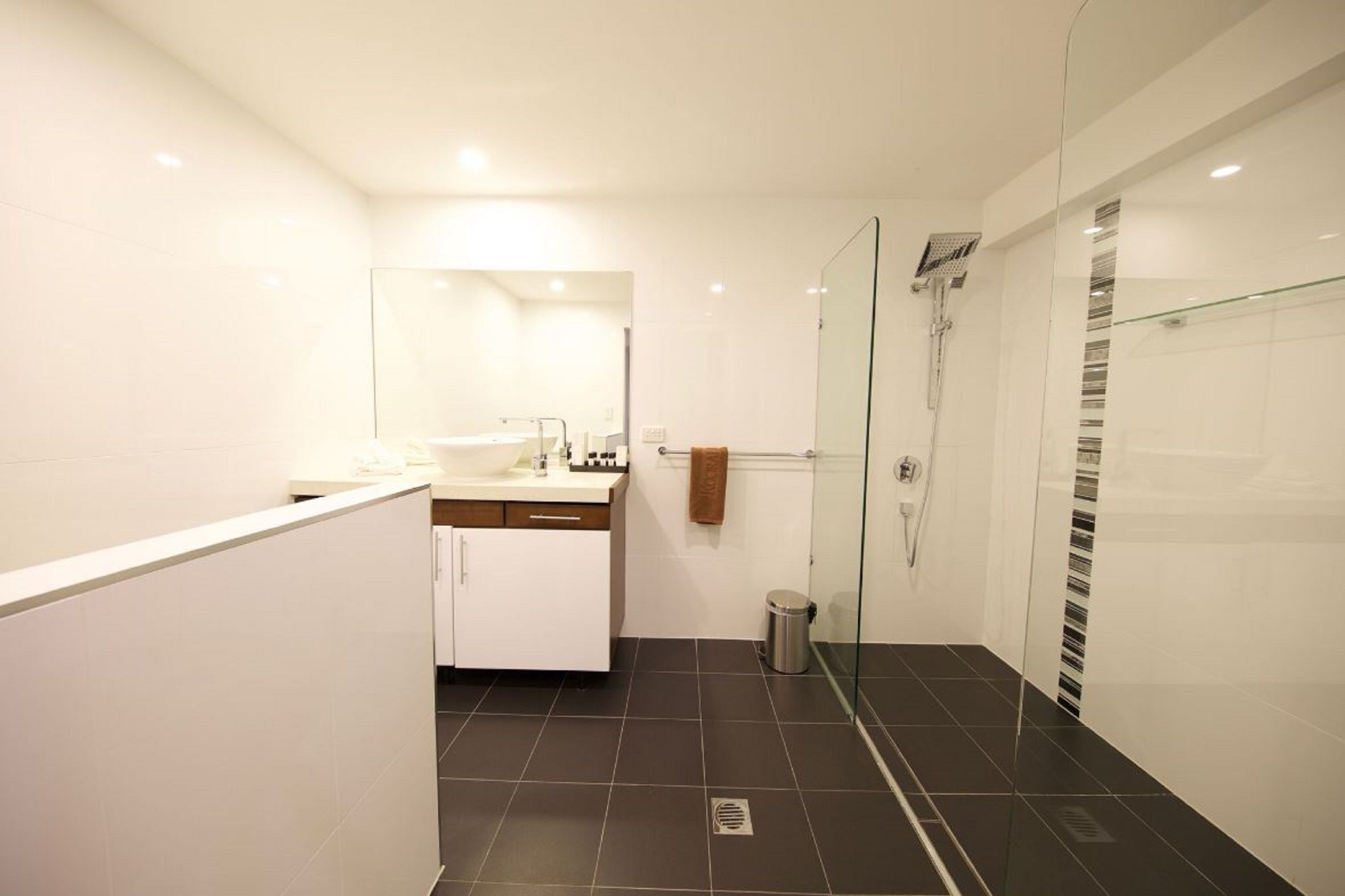 Un baño moderno con cabina de ducha de vidrio, tocador blanco con espejo y suelo de baldosas oscuras.