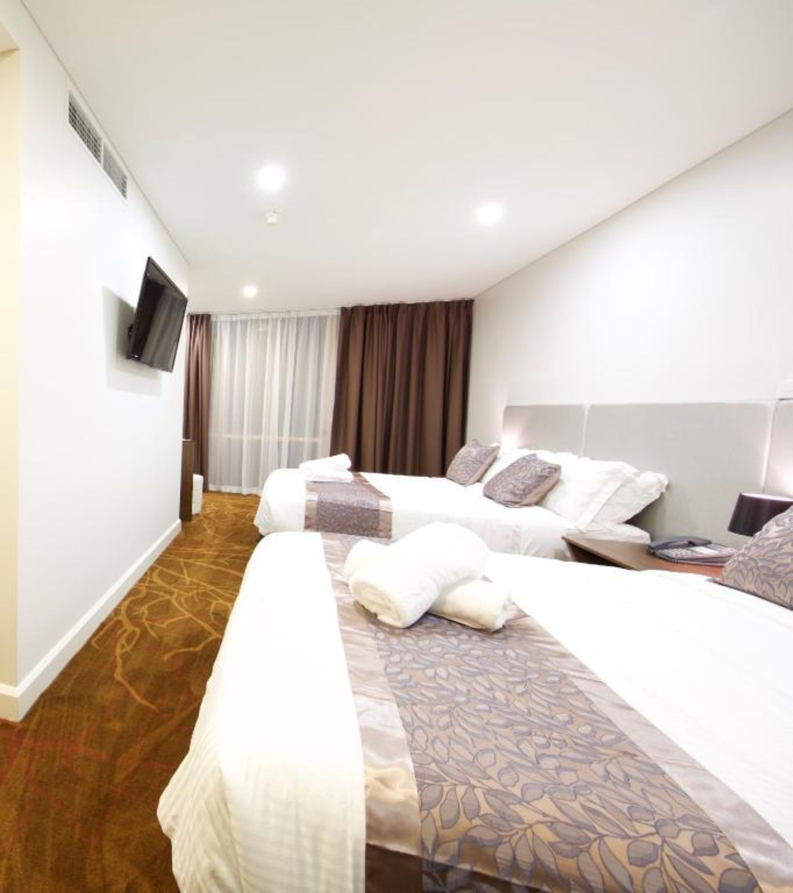 Une chambre d’hôtel avec deux lits, un téléviseur à écran plat fixé au mur, une fenêtre avec des rideaux et un sol recouvert de moquette.