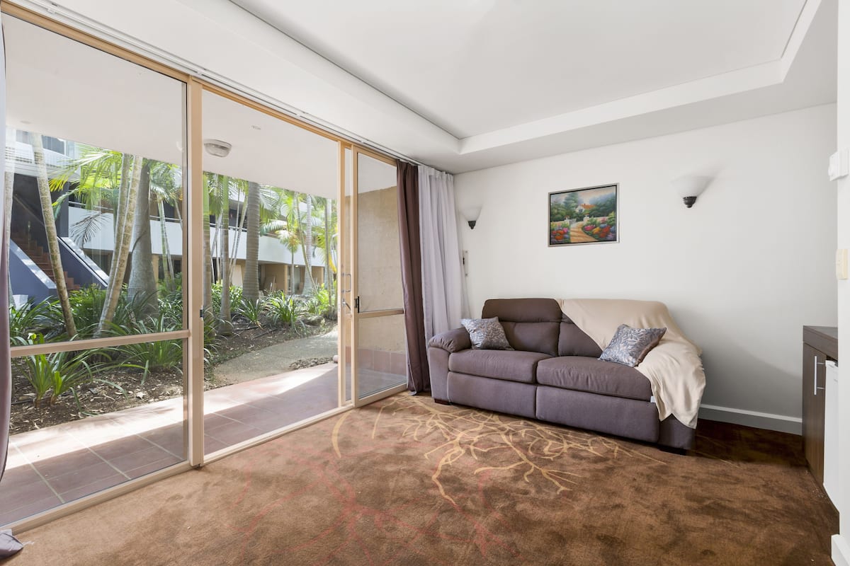 A living room with a sliding glass door leading to a garden, a sofa with cushions, and a painting on the wall.