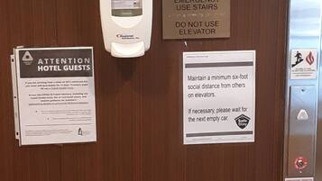 A wooden door with multiple signs, including one advising to use stairs in case of emergency and another about maintaining social distance on elevators.