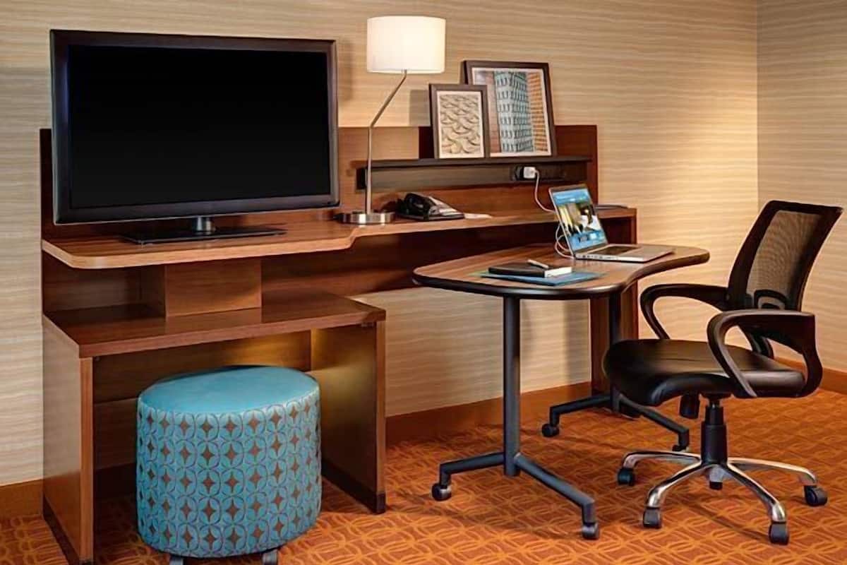A hotel room with a wooden desk, a television, a lamp, a chair, and a patterned ottoman.