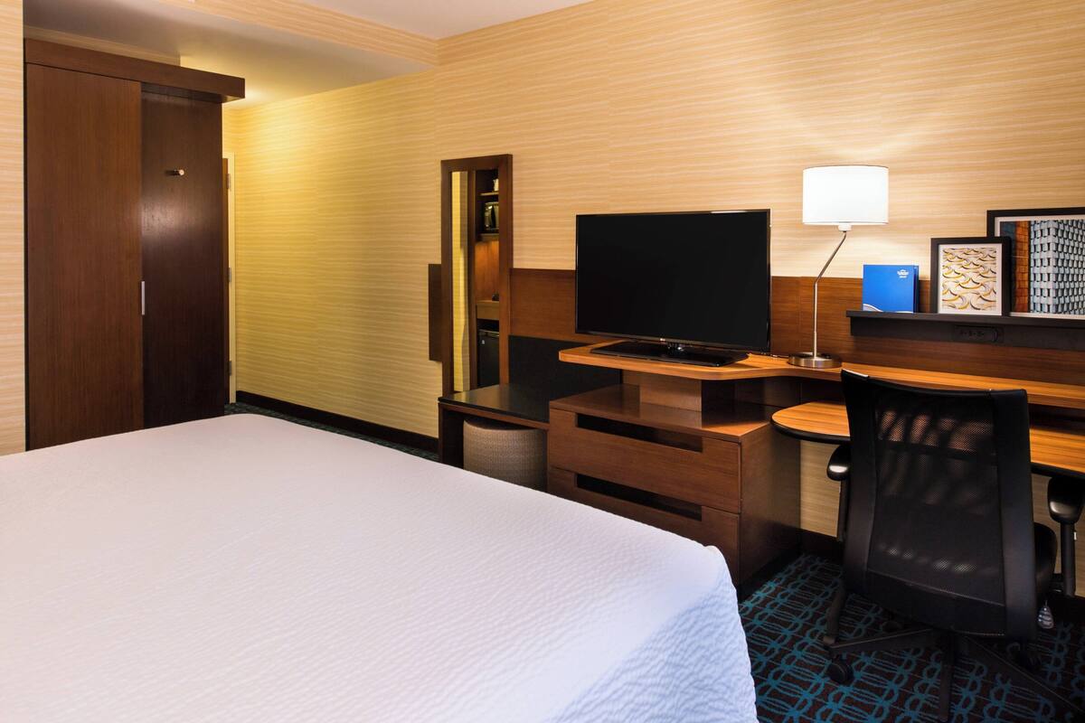 A hotel room with a bed, a desk with a computer, a TV, a chair, and a lamp.