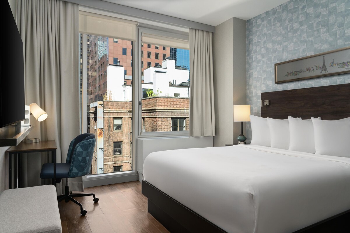 A hotel room with a large bed, a desk with a lamp, a chair, and a view of the cityscape through the window.