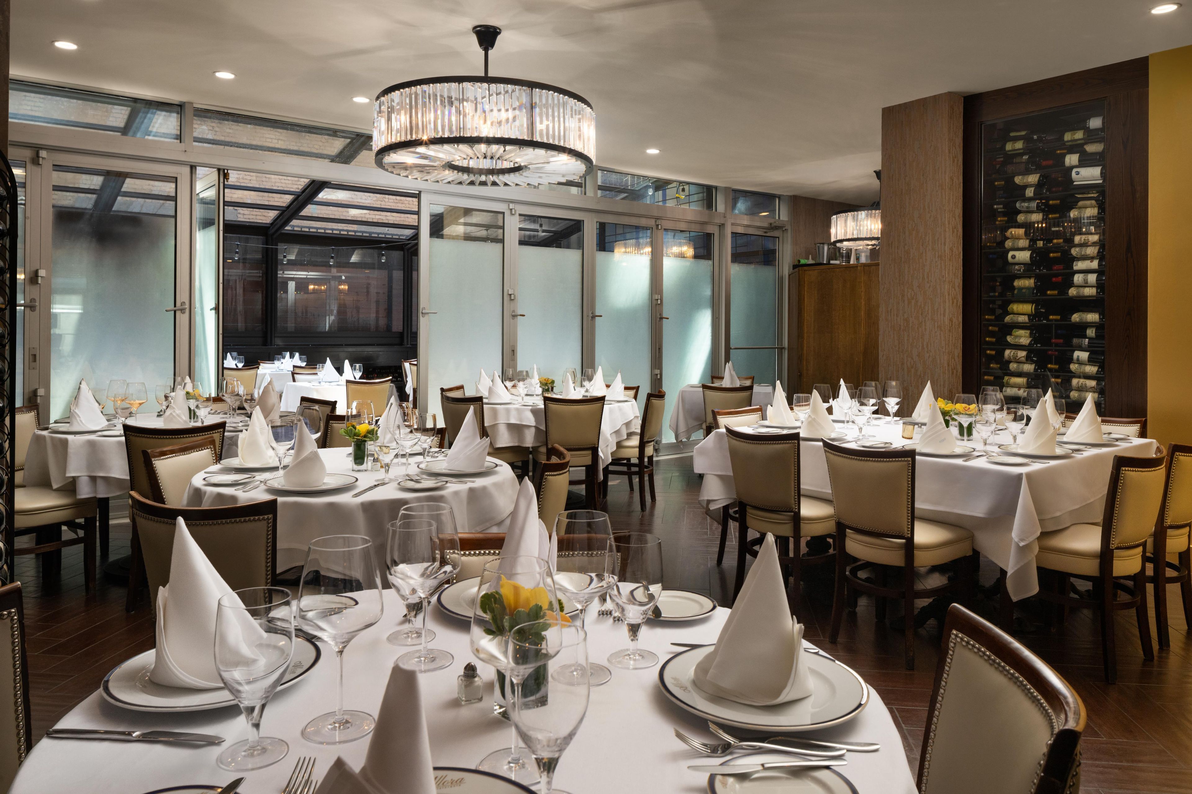 A restaurant interior with round tables set for dining, a chandelier, and a wine rack in the background.