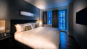 Habitación de hotel con una cama grande, una mesita de noche con teléfono, una ventana con cortinas y un cuadro de un paisaje urbano en la pared.