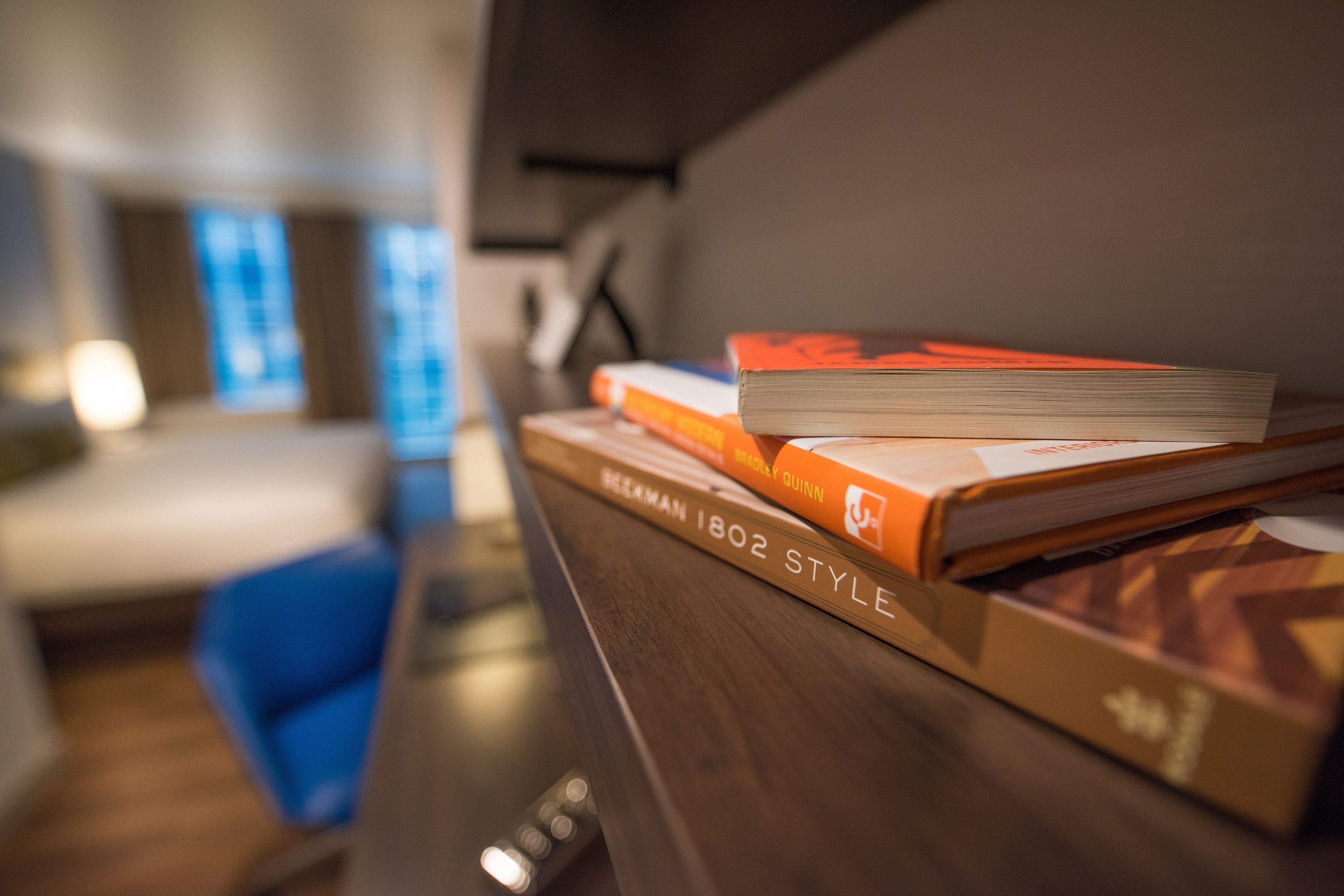 A bookshelf with books, a blue chair, and a wooden desk in a room with a window and a lamp.