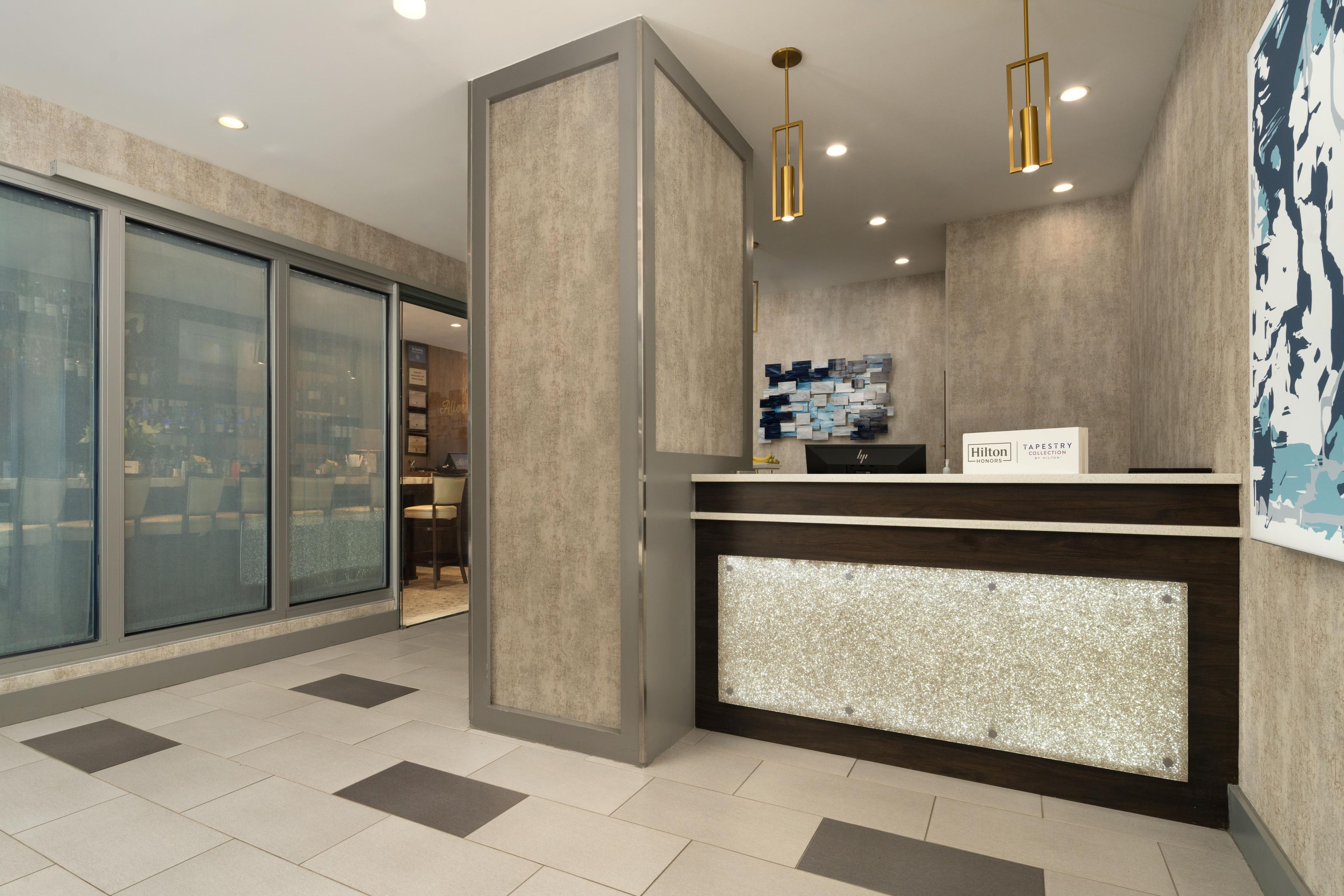 A hotel reception area with a modern design, including a reception desk, a wall-mounted artwork, and a glass door leading to a dining area.
