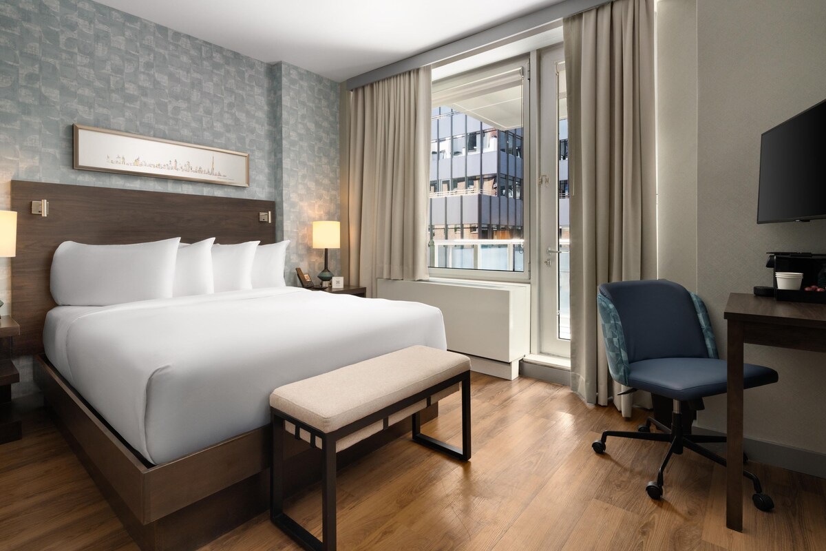 A hotel room with a large bed, bedside lamps, a desk with a chair, a television, and a window with curtains.