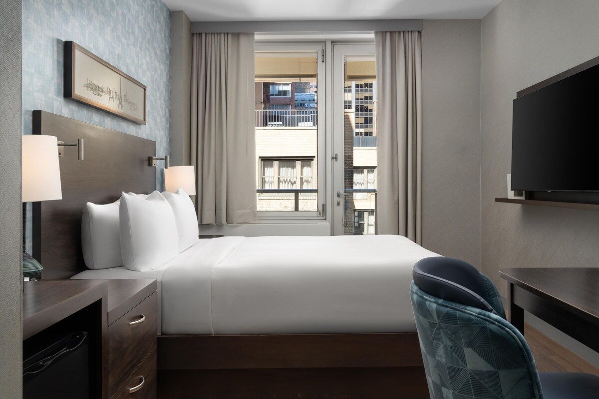 A hotel room with a bed, bedside lamps, a TV, a desk, and a view of the city through the window.