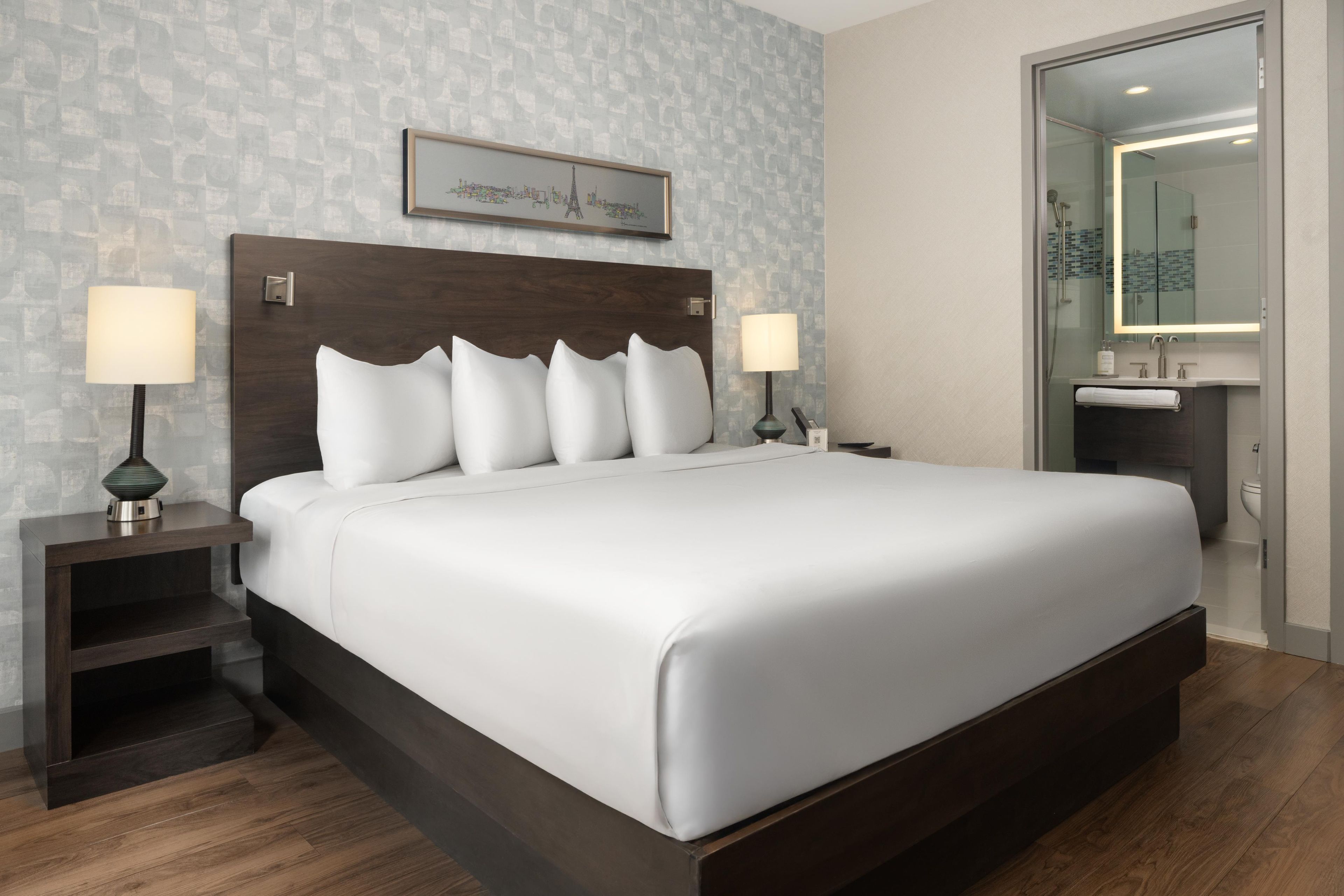 Habitación de hotel con una cama grande, dos mesitas de noche con lámparas, un cuadro enmarcado en la pared y un baño visible a través de una puerta abierta.