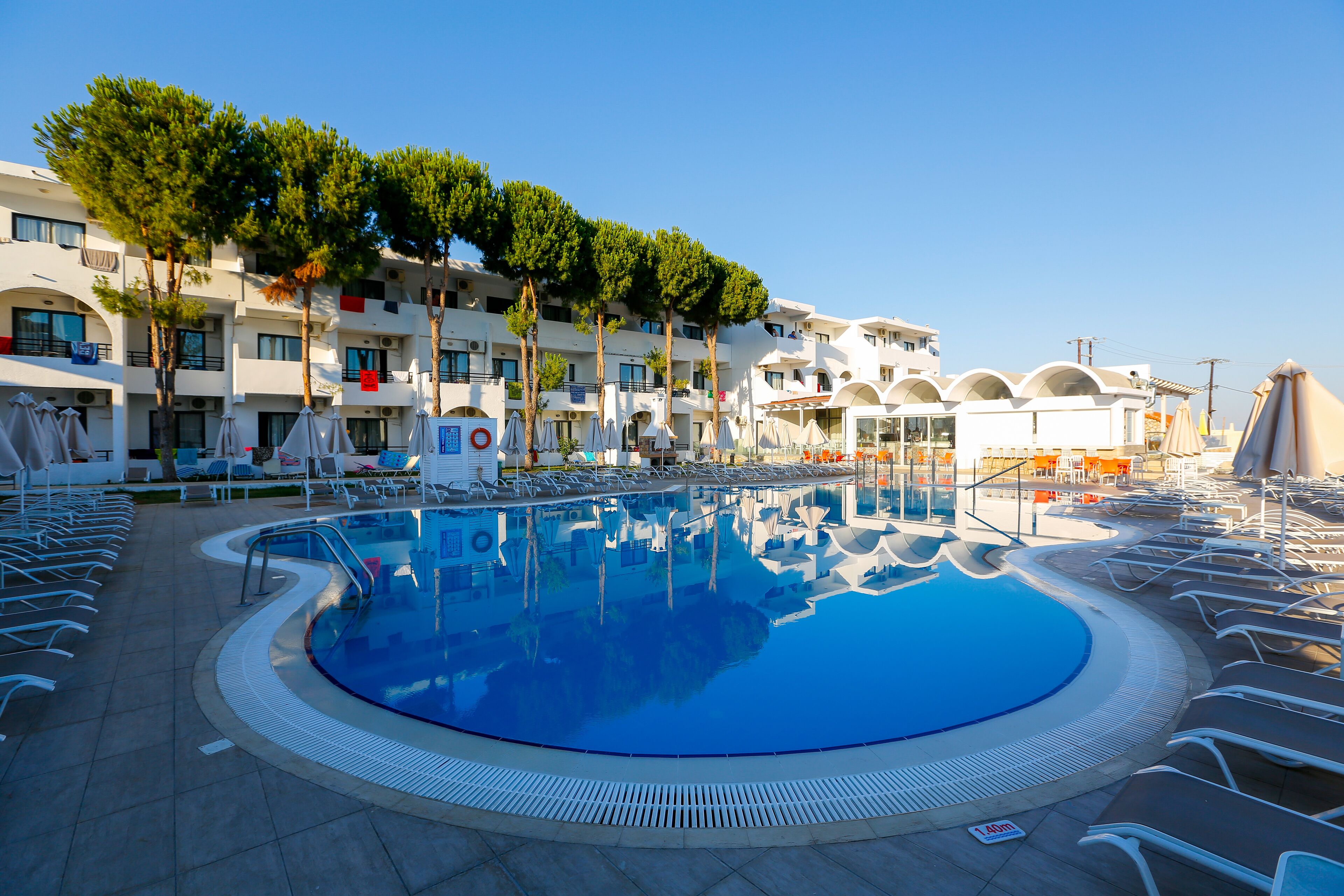 Una piscina con sillones de playa y sombrillas, rodeada por un edificio con balcones y árboles.