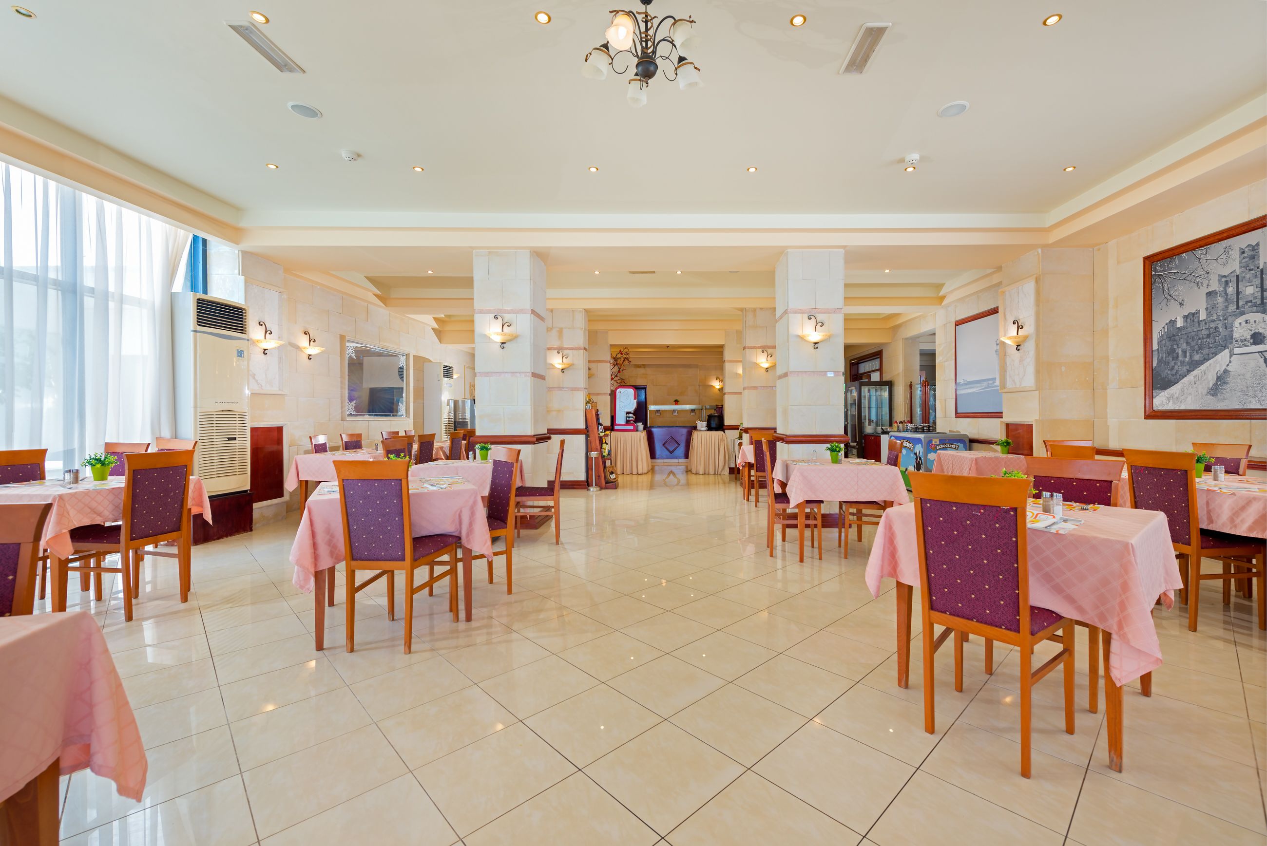 A spacious dining area with multiple tables and chairs, a chandelier, and a large window providing natural light.