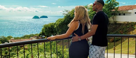 A couple standing on a balcony overlooking a scenic view of the sea and greenery.
