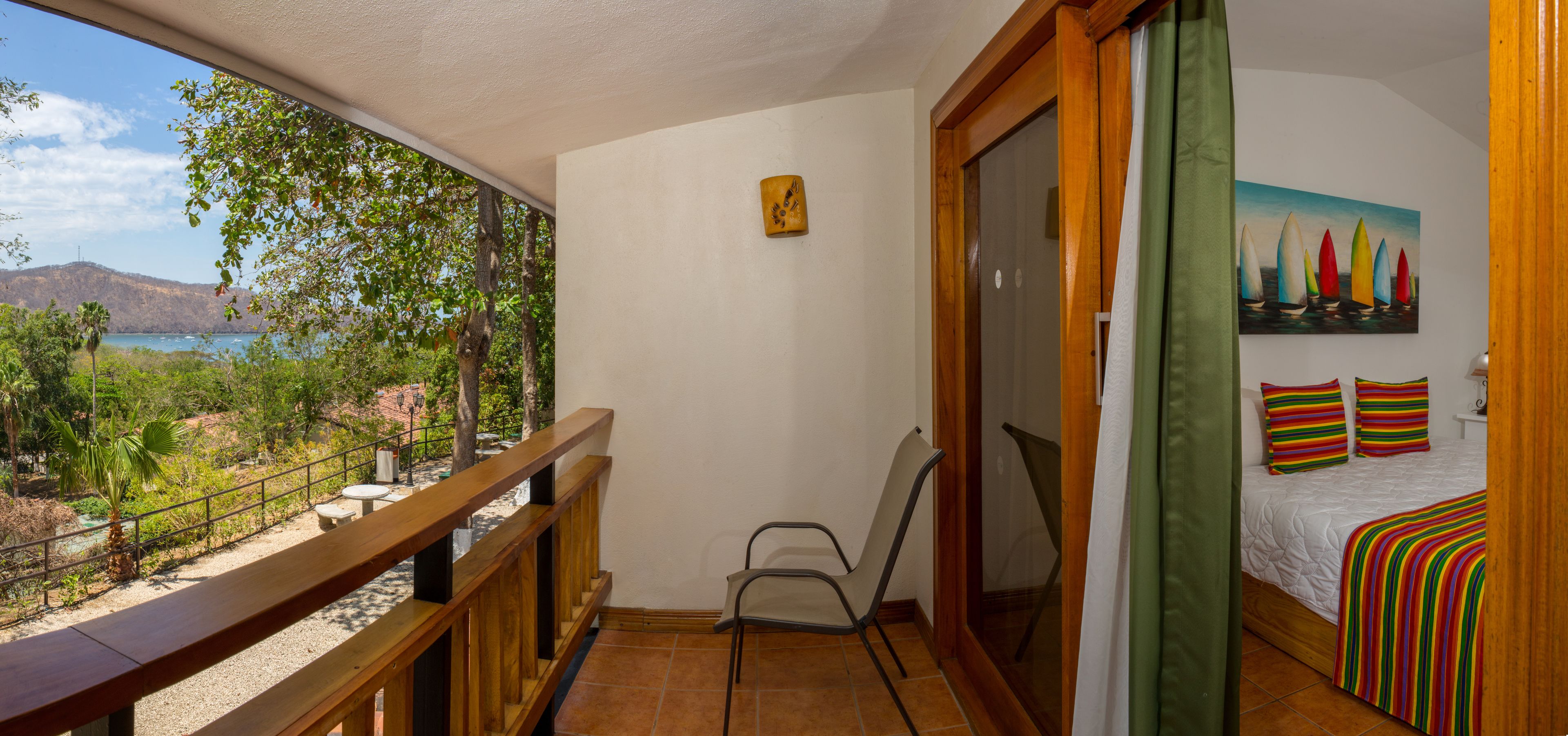 A balcony with a wooden railing, a chair, a bed with colorful pillows, and a painting of sailboats.