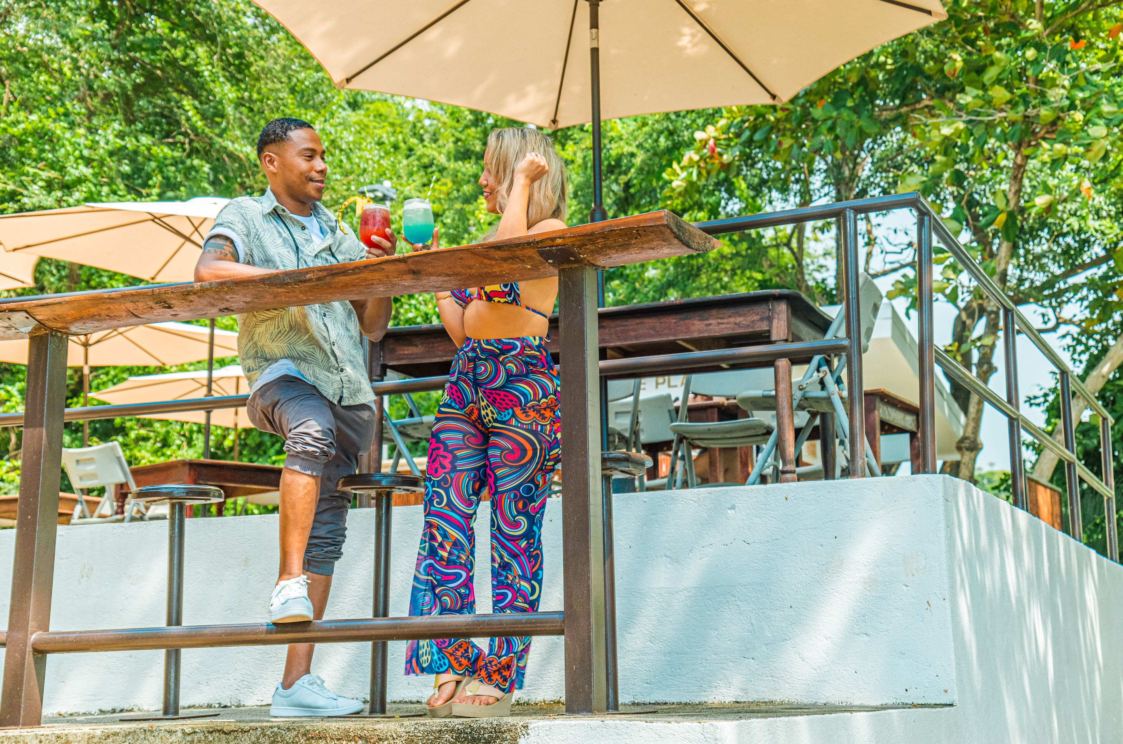 Un hombre y una mujer sentados en una mesa al aire libre, con una bebida, rodeados de follaje y una baranda blanca.