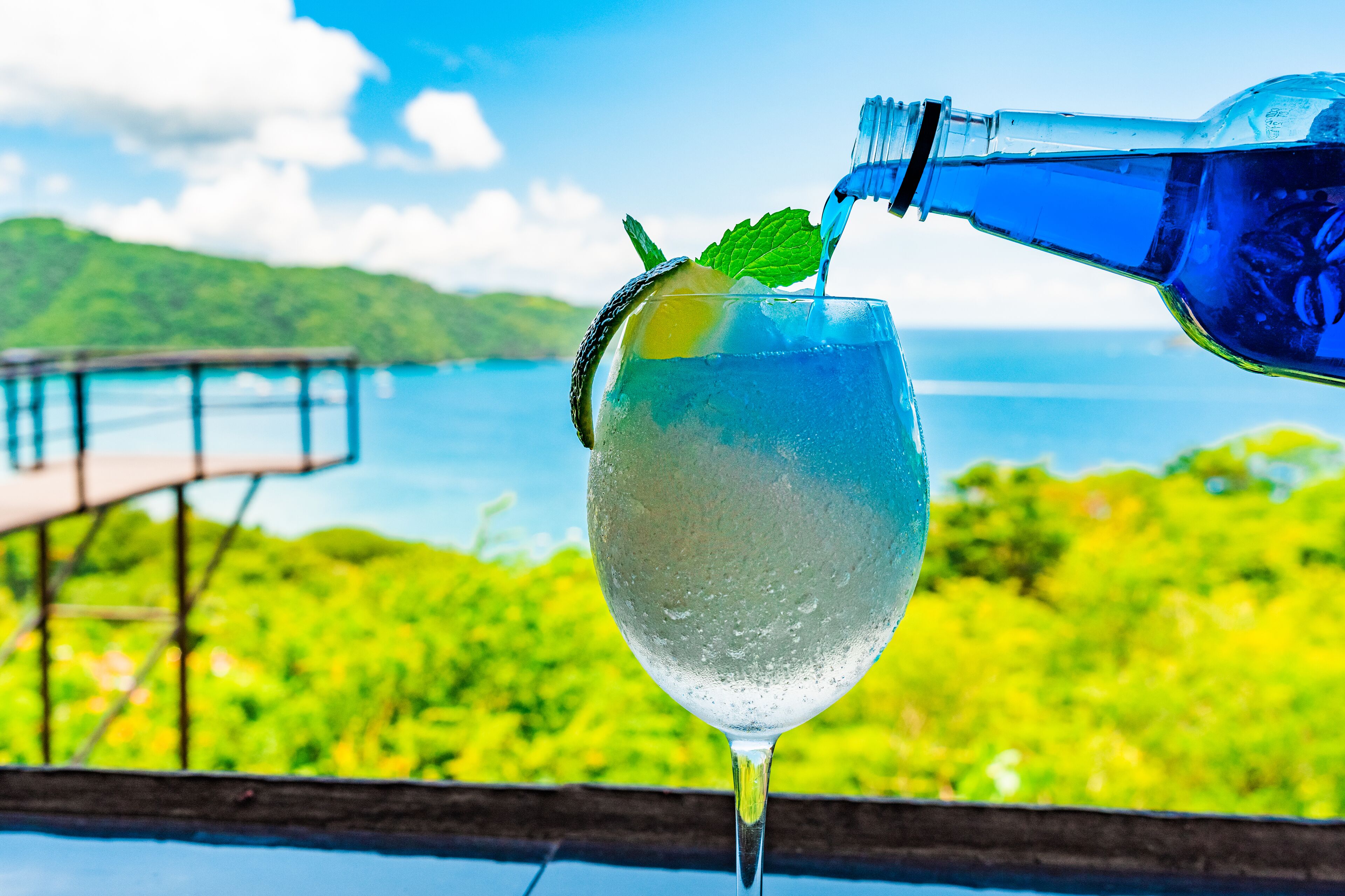 A glass of water with mint and lemon, with a scenic view of a bridge and greenery in the background.