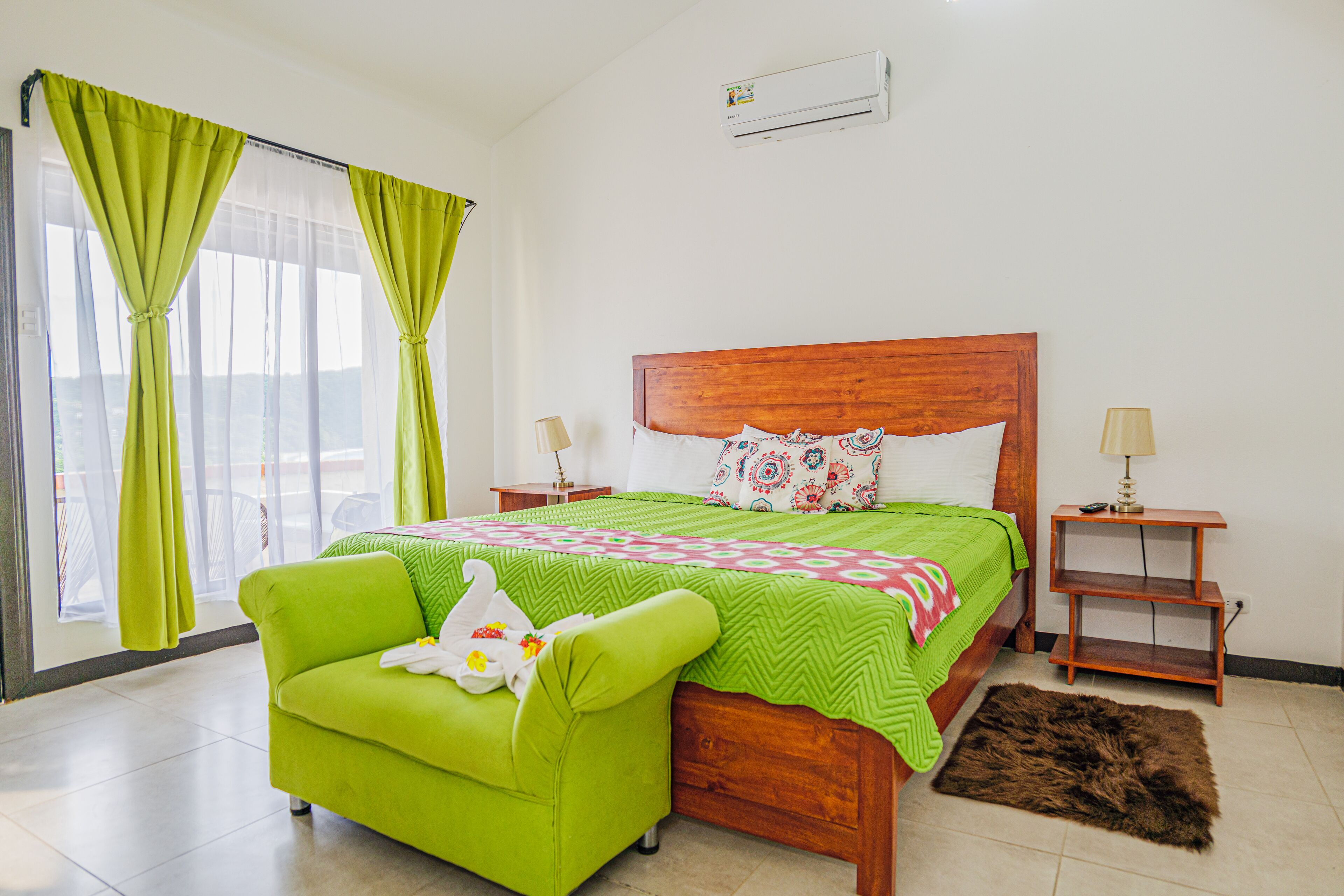 Un dormitorio con una cama de madera, cortinas verdes, un sofá verde, una alfombra pequeña y dos mesitas de noche con lámparas.