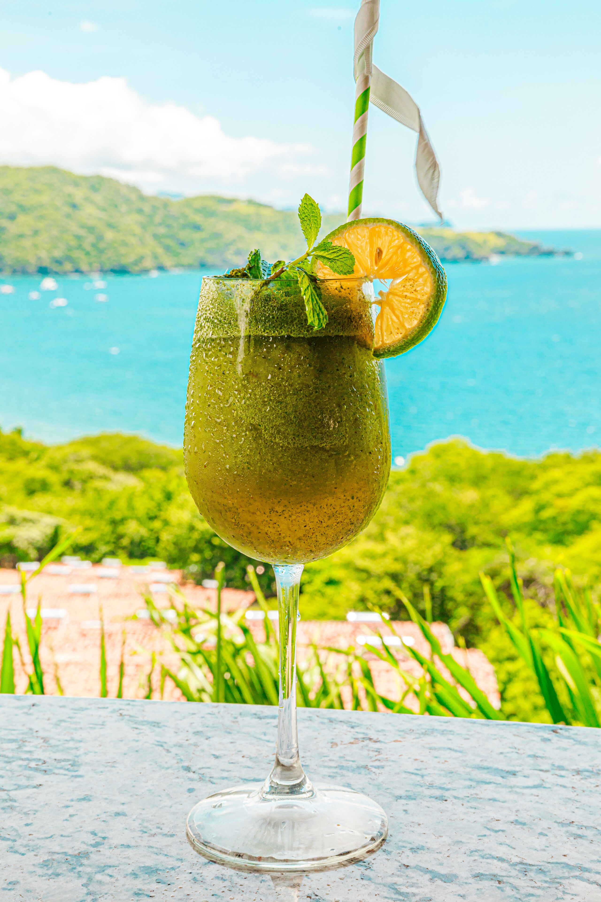A glass of drink with a slice of orange and mint on a table with a view of the sea and hills in the background.