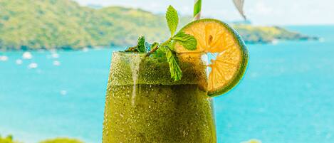 A glass of drink with a slice of orange and mint on a table with a view of the sea and hills in the background.
