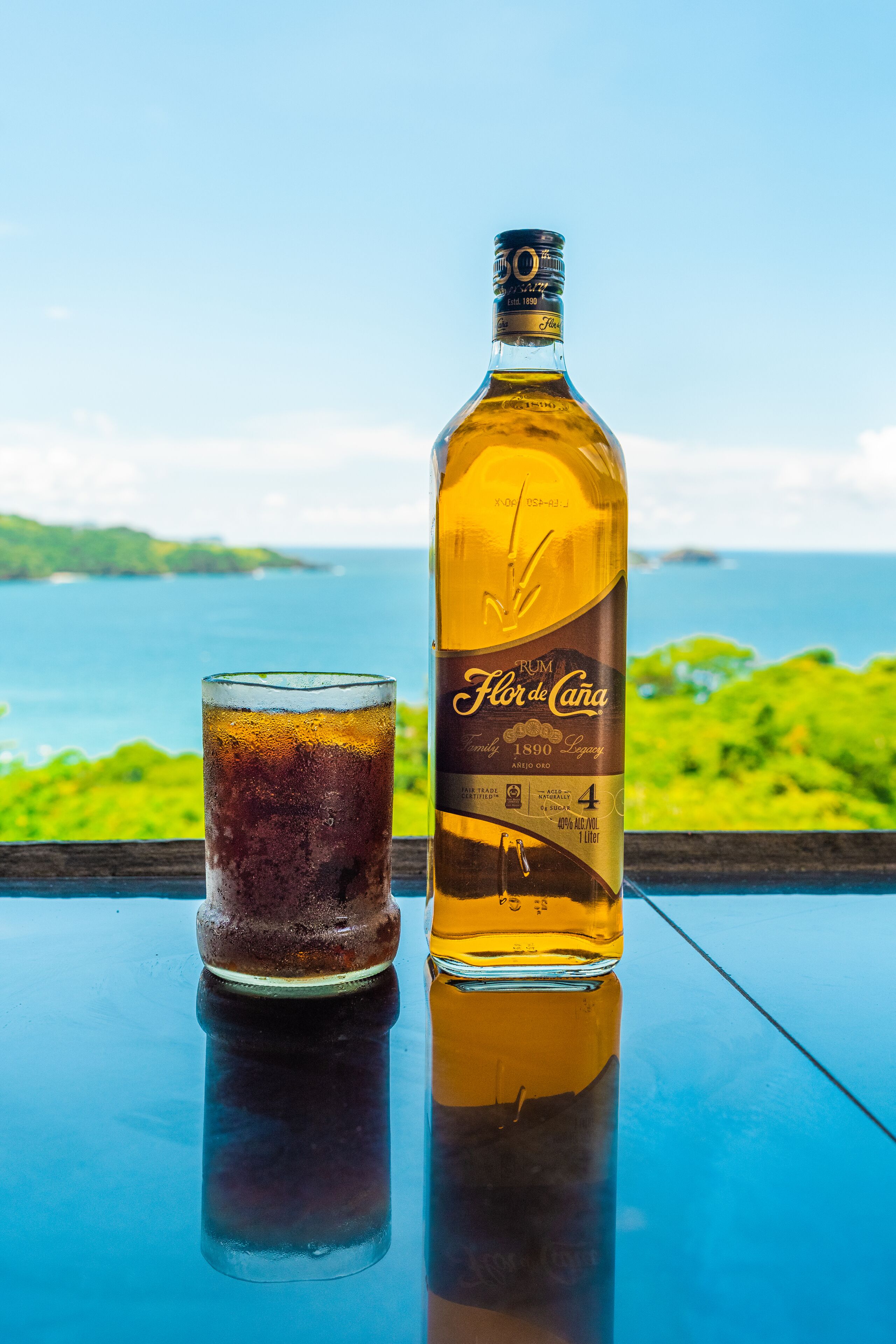 A bottle of Flor de Cana rum and a glass of dark liquid on a table with a view of the sea.