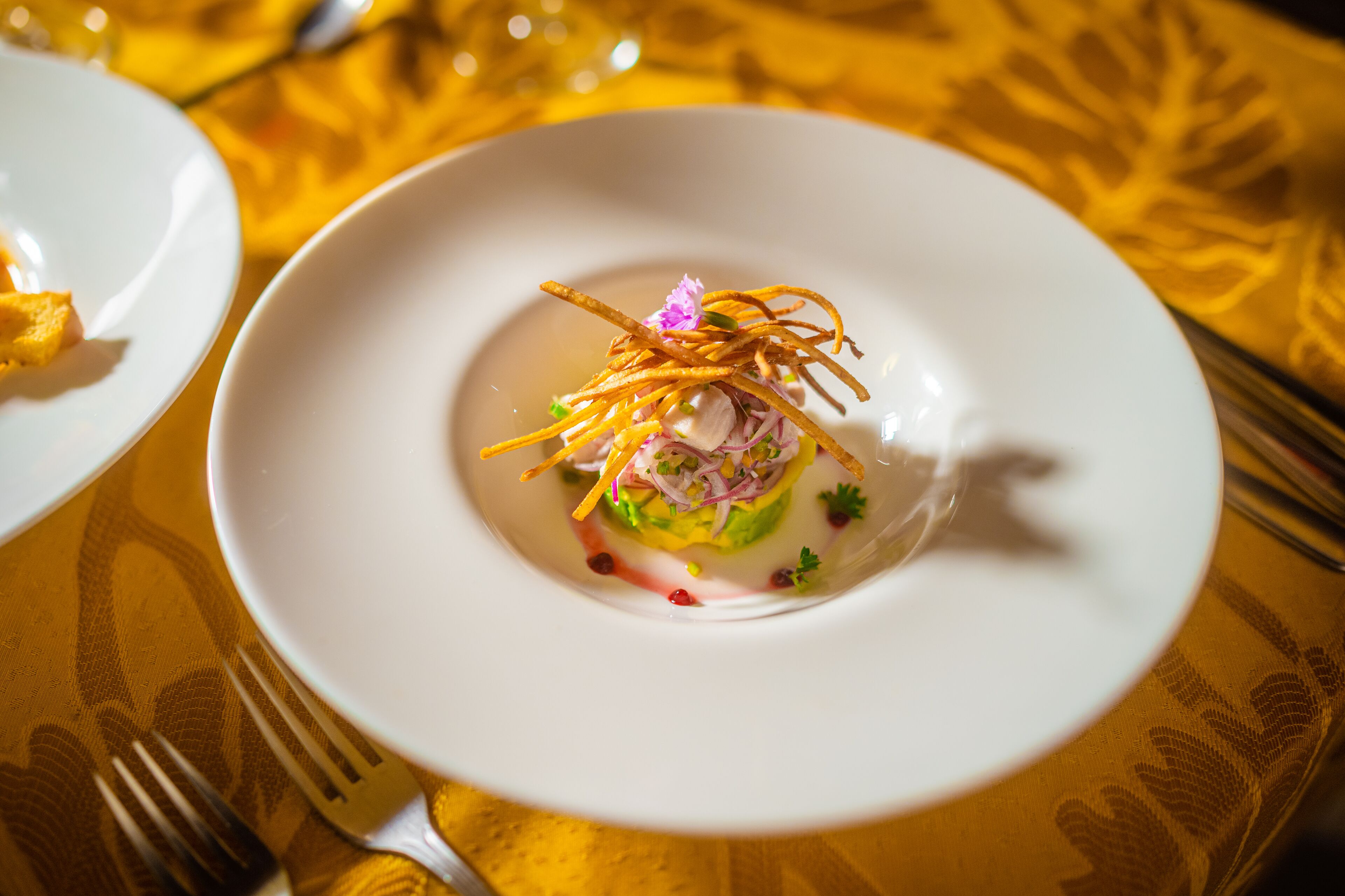 A white plate with a colorful dish, possibly a salad, garnished with thin strips and a flower, on a table with a yellow patterned tablecloth.