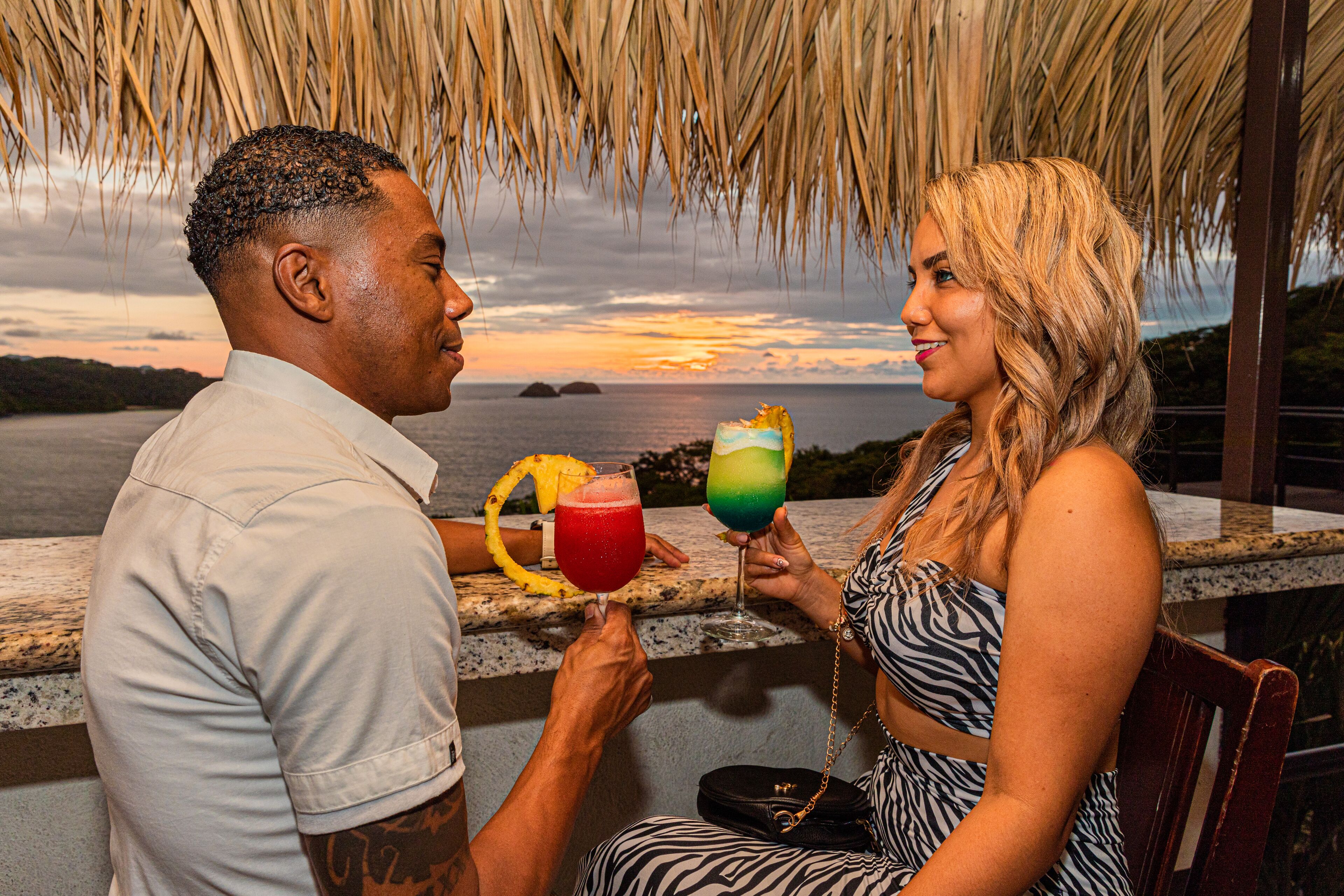 Un couple est assis à une table d’où l’on a une vue sur l’océan et le coucher de soleil. Ils tiennent chacun un verre décoré de fruits tropicaux.