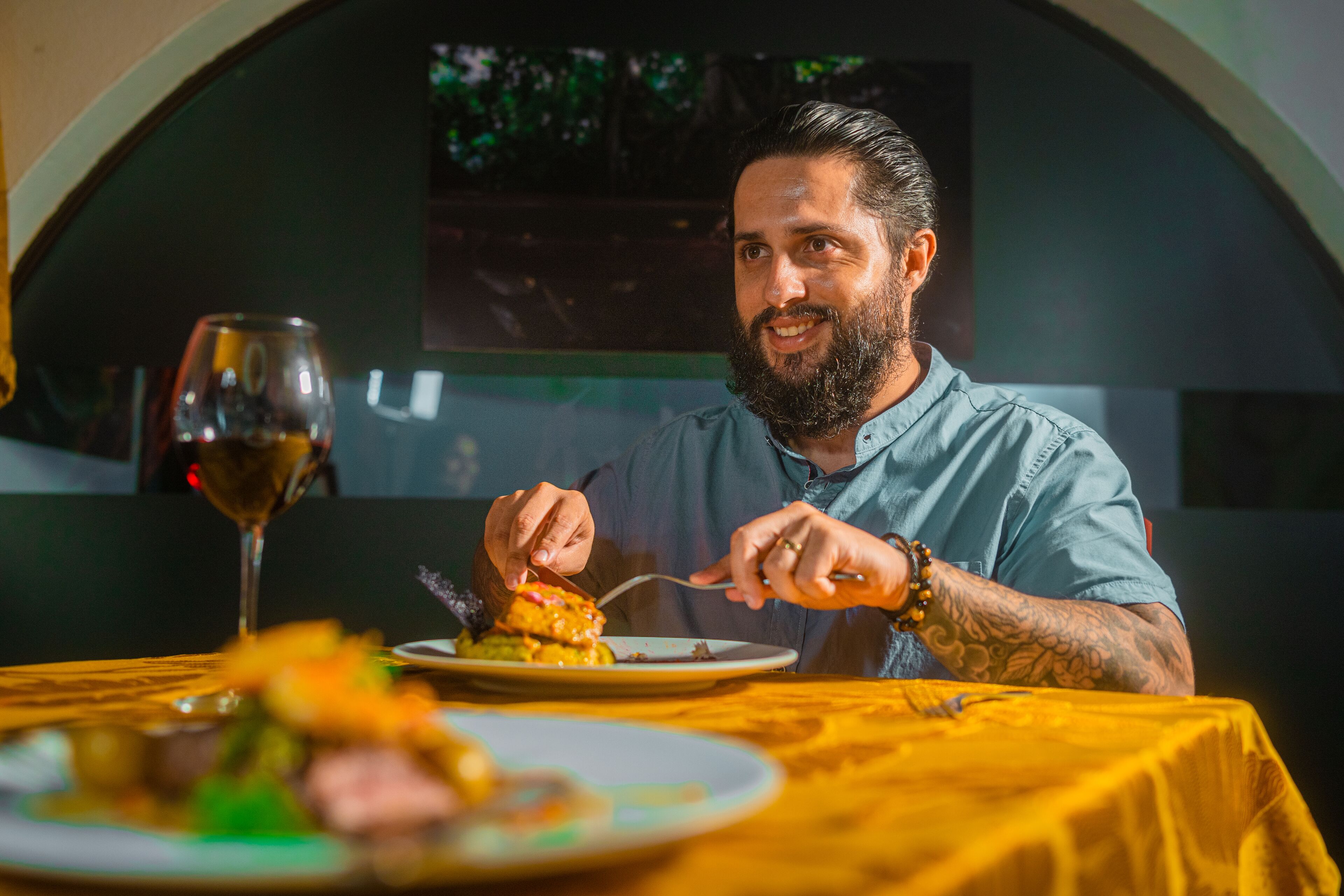 Iemand zit aan een tafel met een geel tafelkleed, een wijnglas en borden met eten.