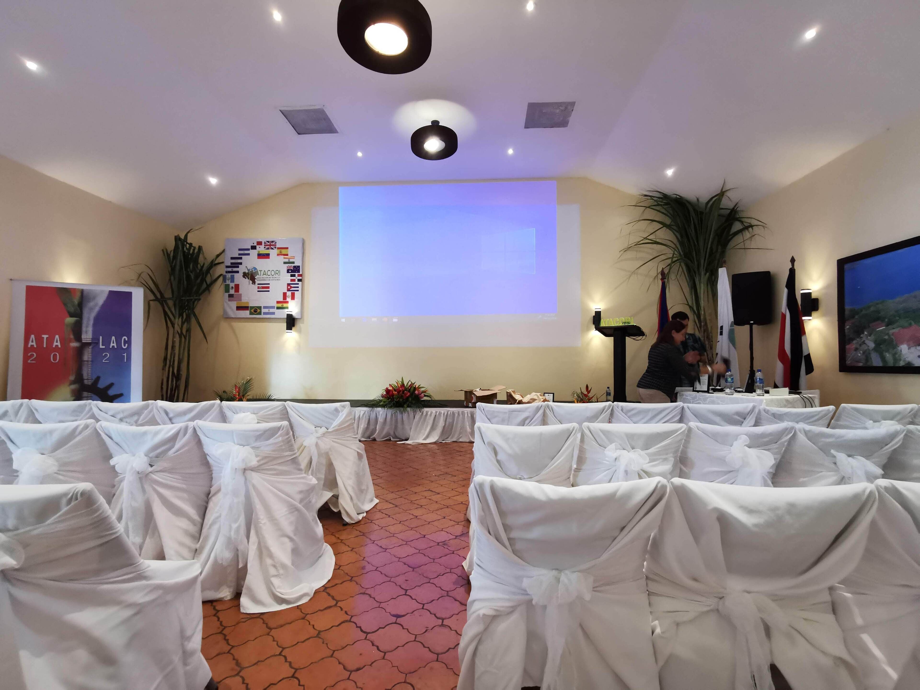 A conference room with rows of white-covered chairs, a projector screen, and a podium with a microphone.