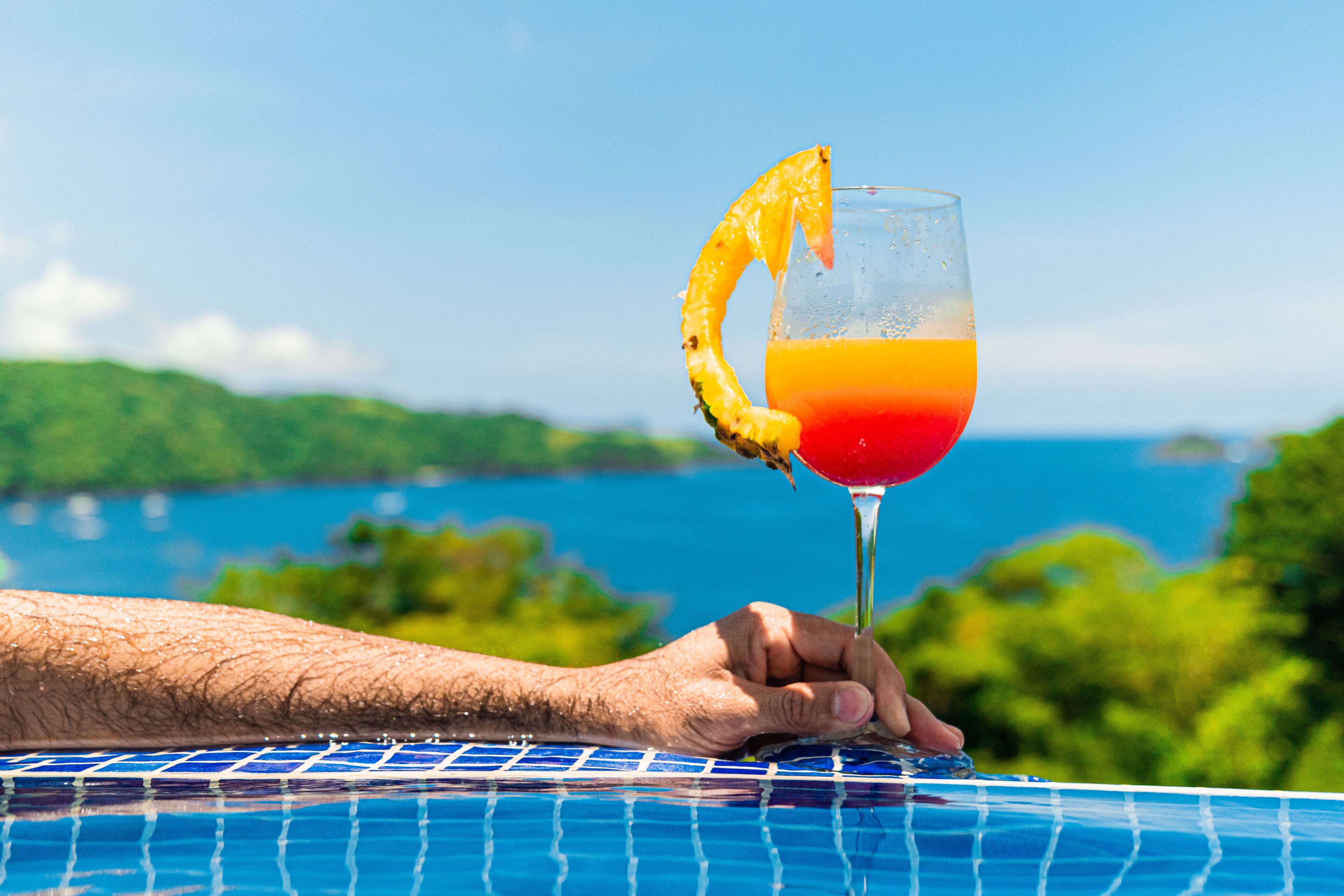 A hand holding a cocktail glass with a slice of pineapple on the rim, set against a scenic view of a body of water and greenery.