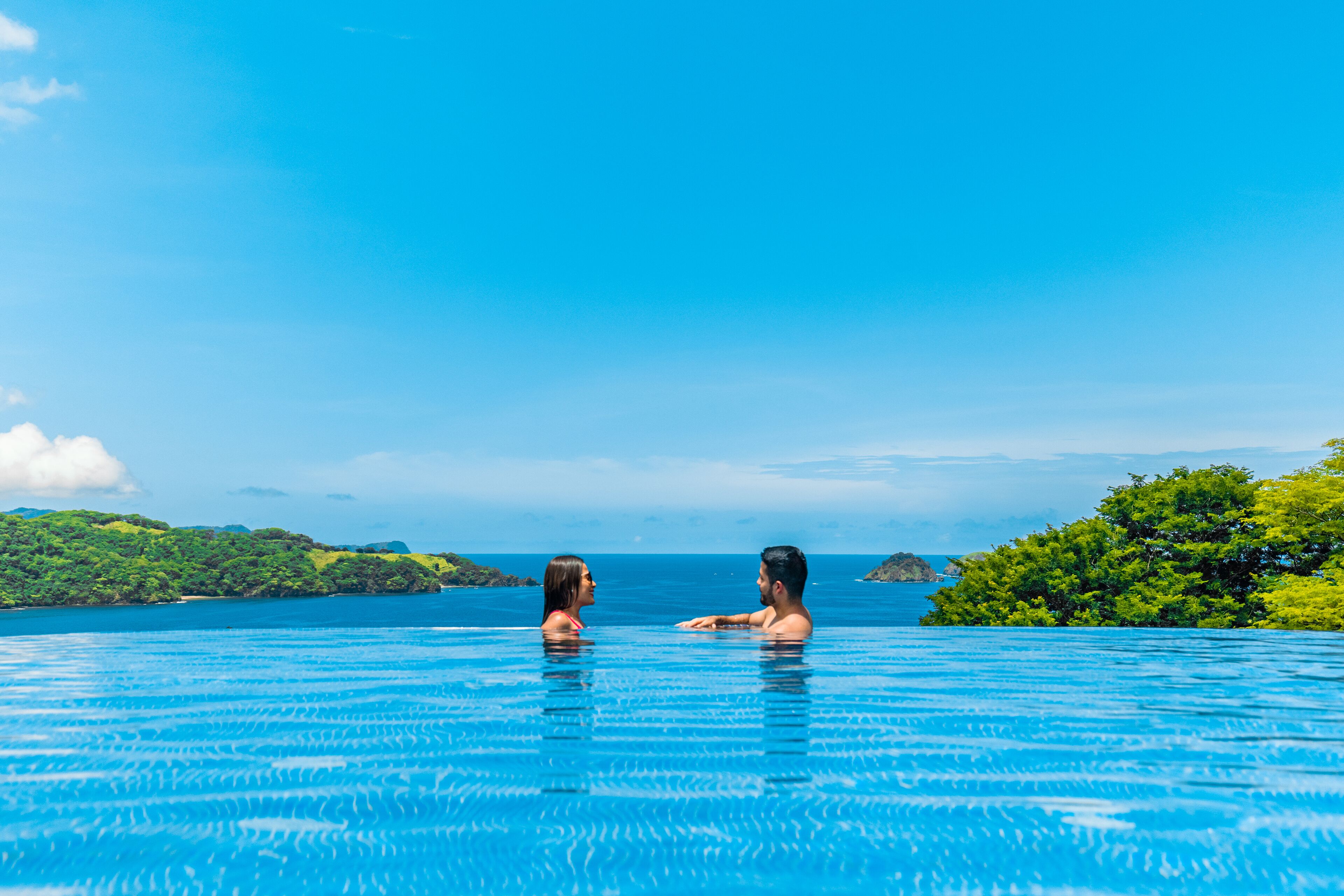 Un couple se détend dans une piscine à débordement aux eaux d’un bleu limpide, avec vue sur l’océan et la végétation luxuriante.