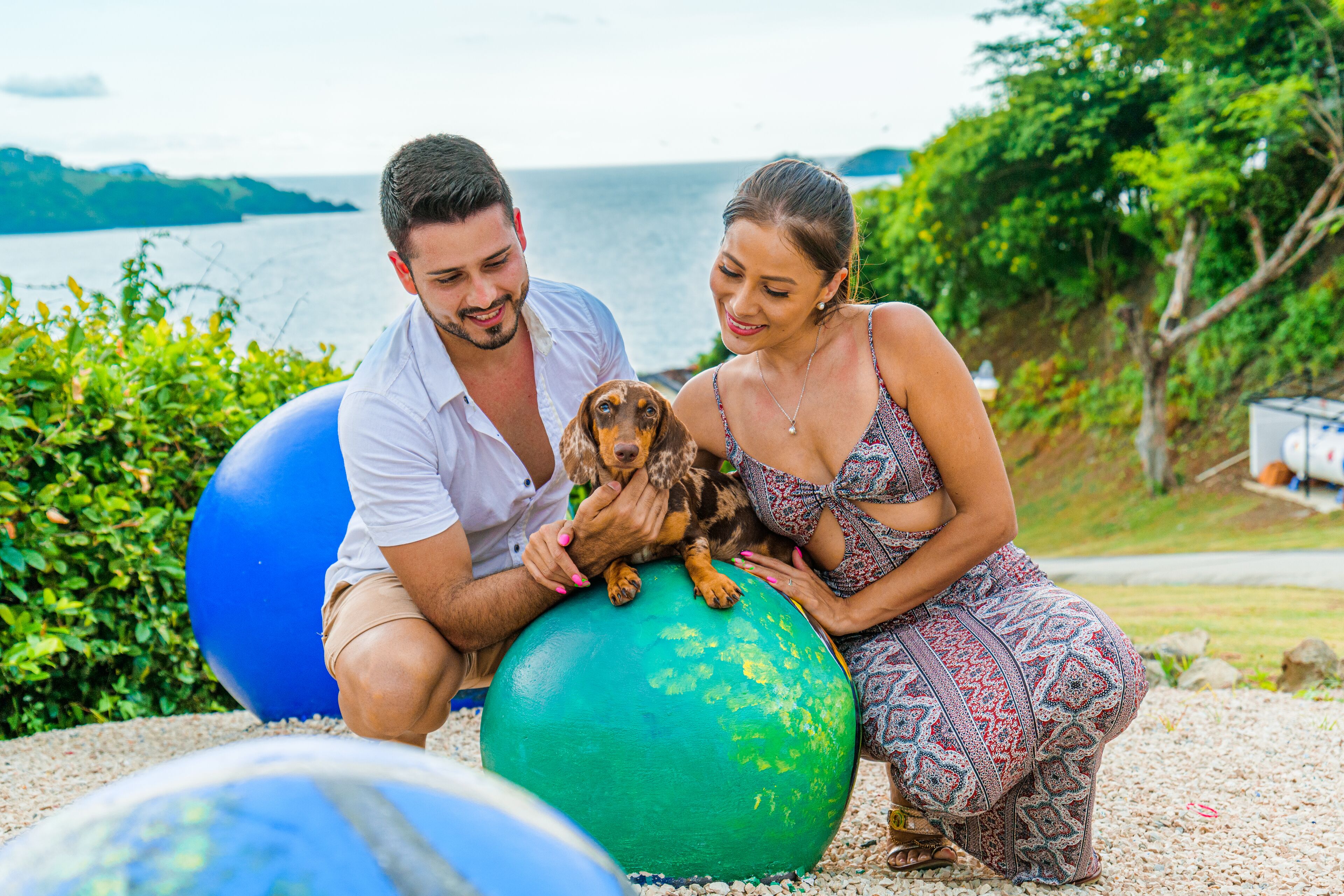 A couple sitting on a large blue exercise ball outdoors with a dog, surrounded by greenery and a body of water in the background.