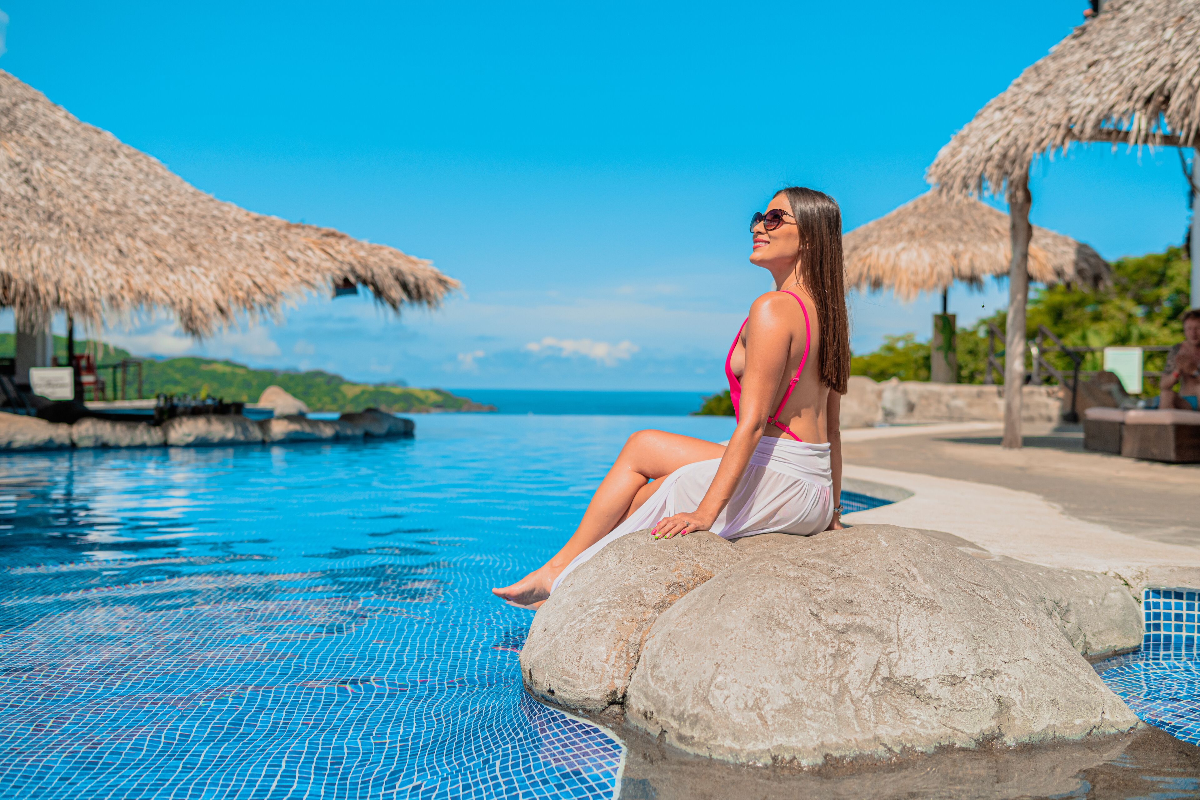 A woman sitting on a rock by a pool with a clear blue sky and ocean in the background.