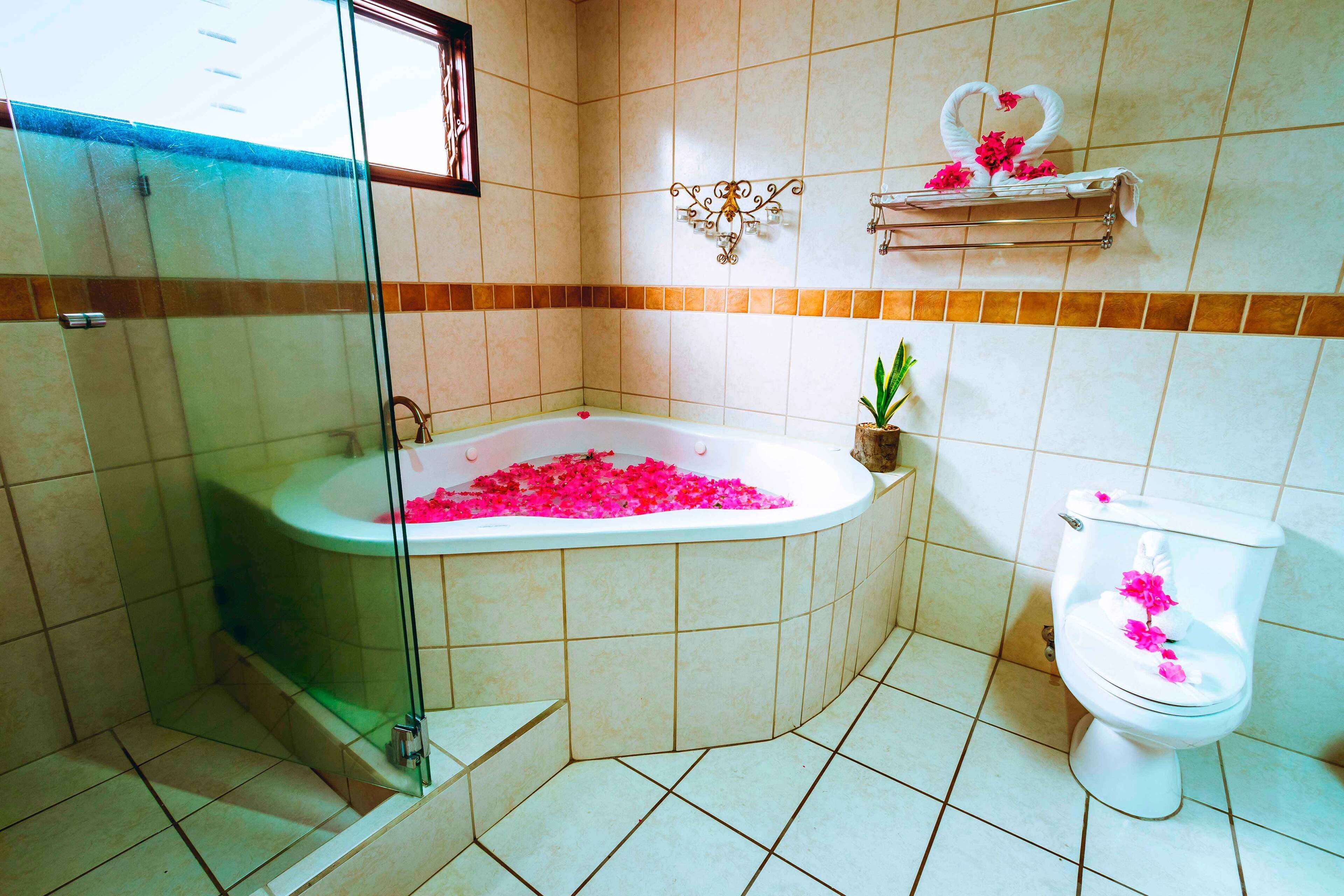 A bathroom with a glass-enclosed shower, a bathtub filled with pink flowers, a toilet decorated with pink flowers, and a small potted plant.
