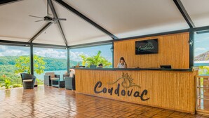 A reception area with a wooden reception desk labeled "Coudovac," a ceiling fan, and a view of lush greenery and hills. - Condovac la Costa (Sardinal)