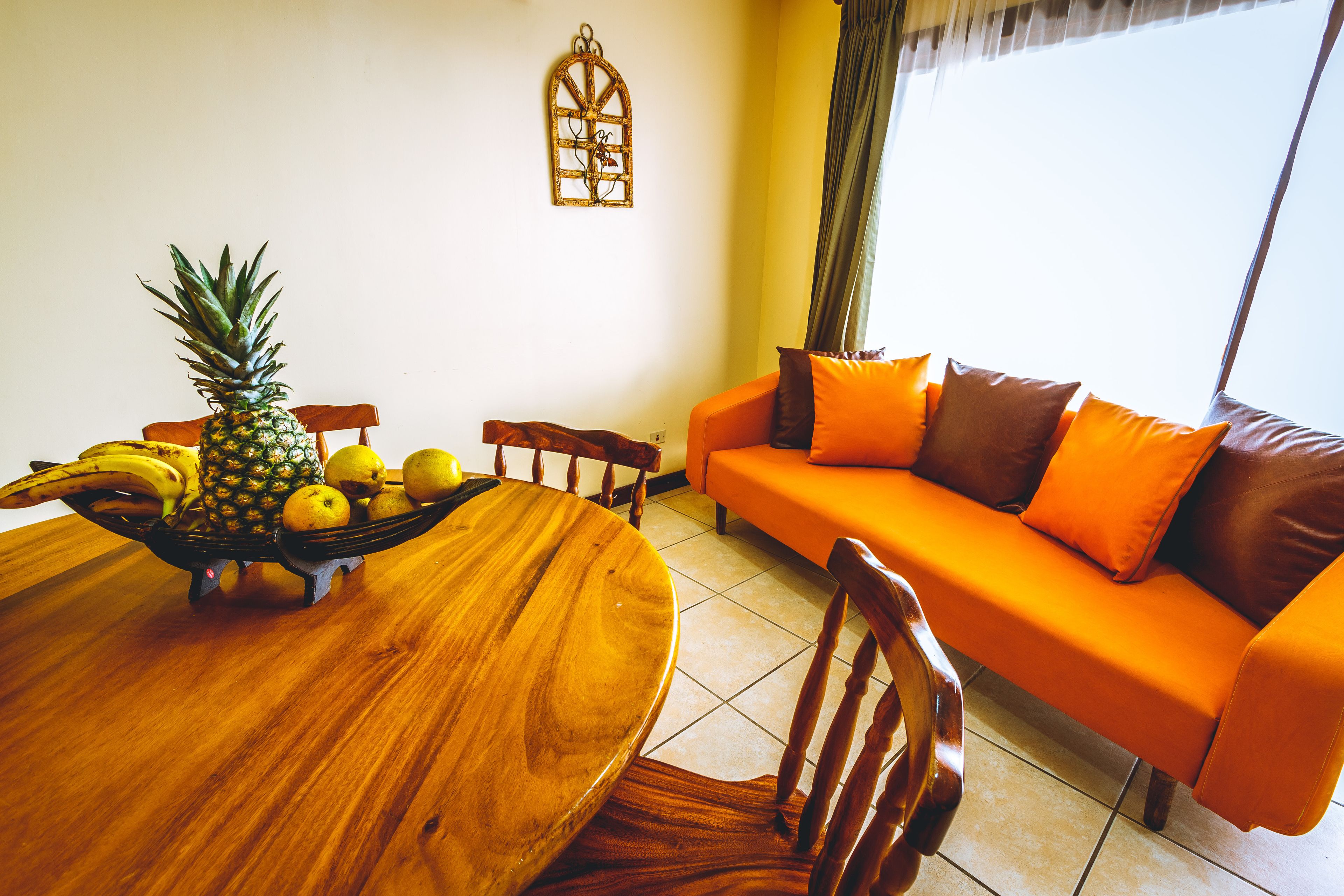 A dining area with a wooden table, chairs, and a fruit bowl containing a pineapple, bananas, and lemons. There is an orange sofa with brown cushions in the background.