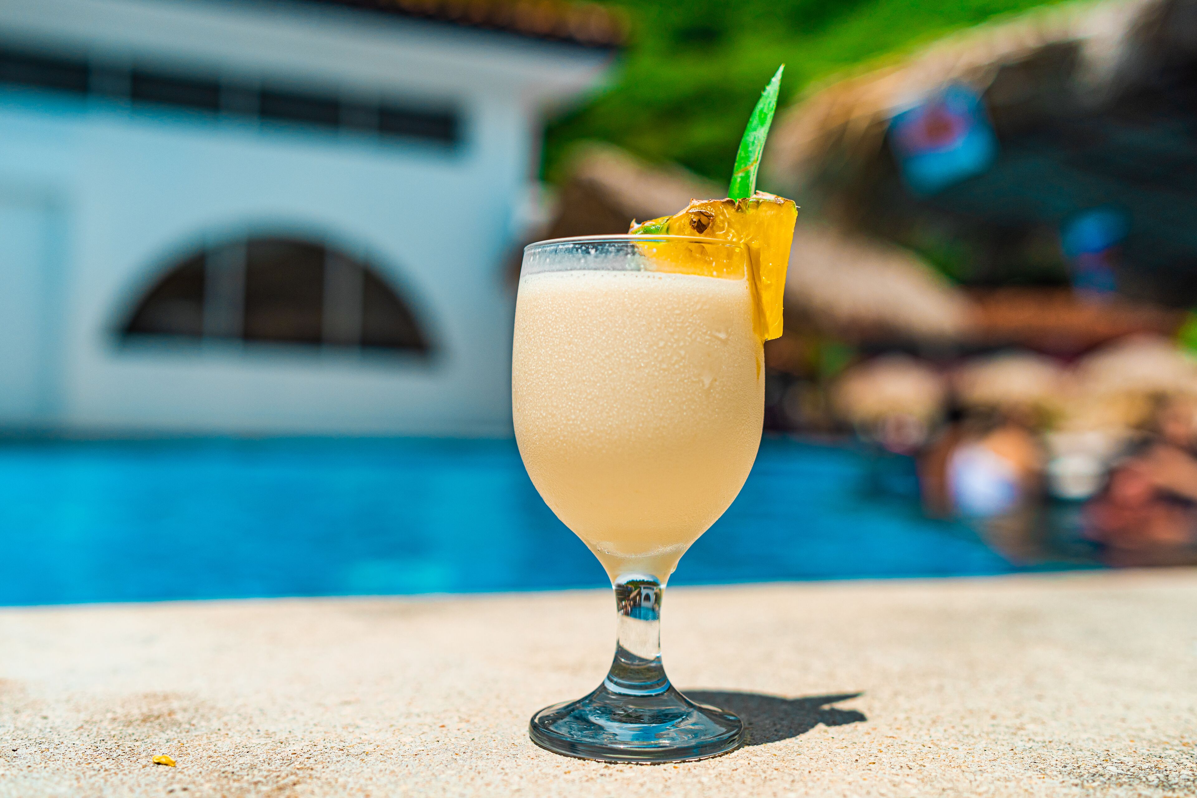 A cocktail glass with a straw and a slice of fruit on a sandy surface near a pool.