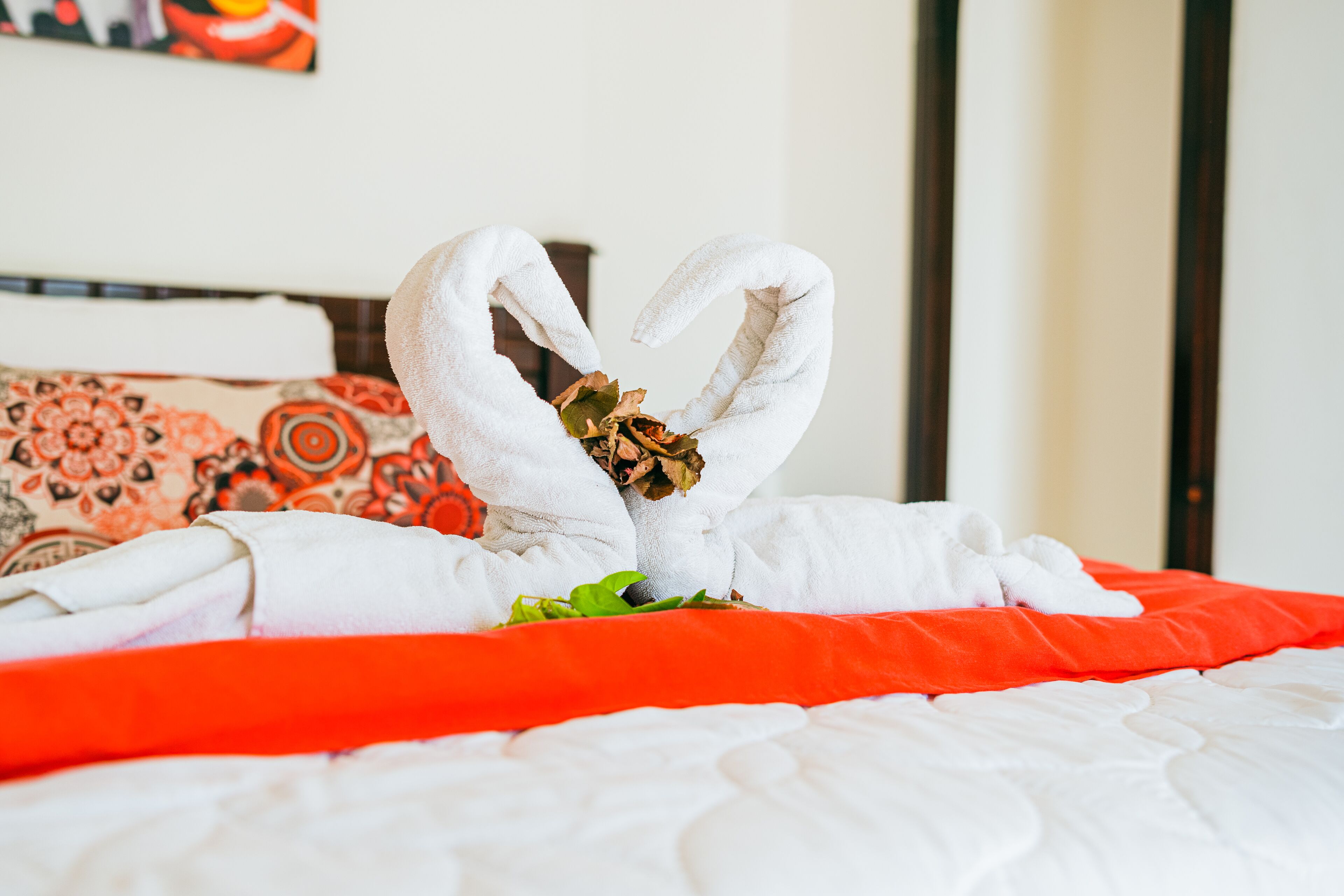 A bed with a red blanket and a white quilt, a heart-shaped towel arrangement, and a decorative pillow.