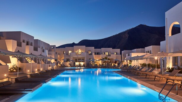 A swimming pool with lounge chairs and umbrellas, surrounded by buildings with a mountainous backdrop. - Aqua Blue Hotel (Santorini)