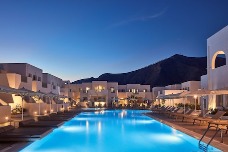A swimming pool with lounge chairs and umbrellas, surrounded by buildings with a mountainous backdrop.