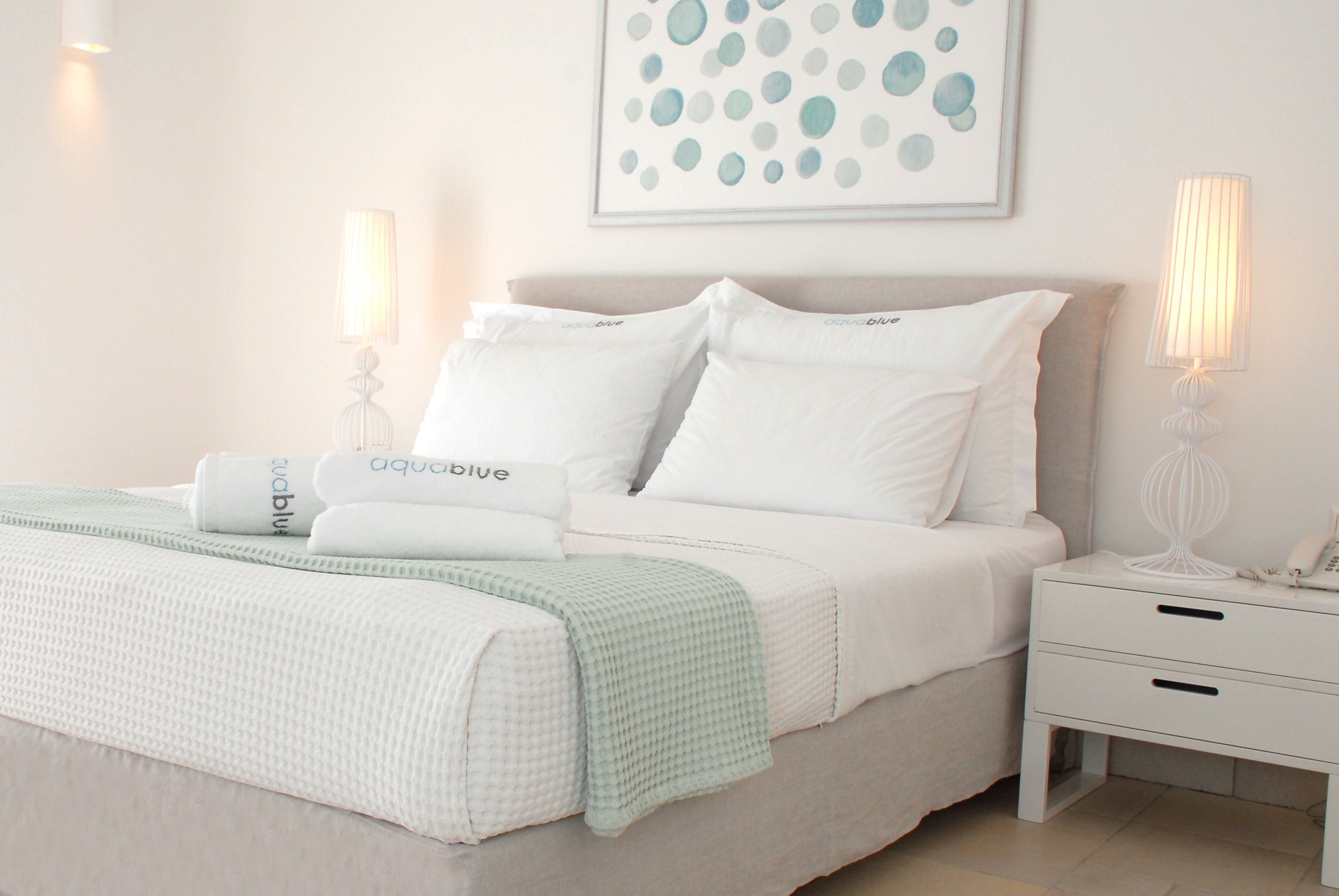 A neatly made bed with white bedding and a green checkered throw, a bedside table with a lamp, and a wall-mounted artwork.