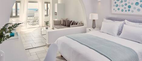 A modern hotel room with a large bed, a desk, and a view of the sea through an arched doorway.