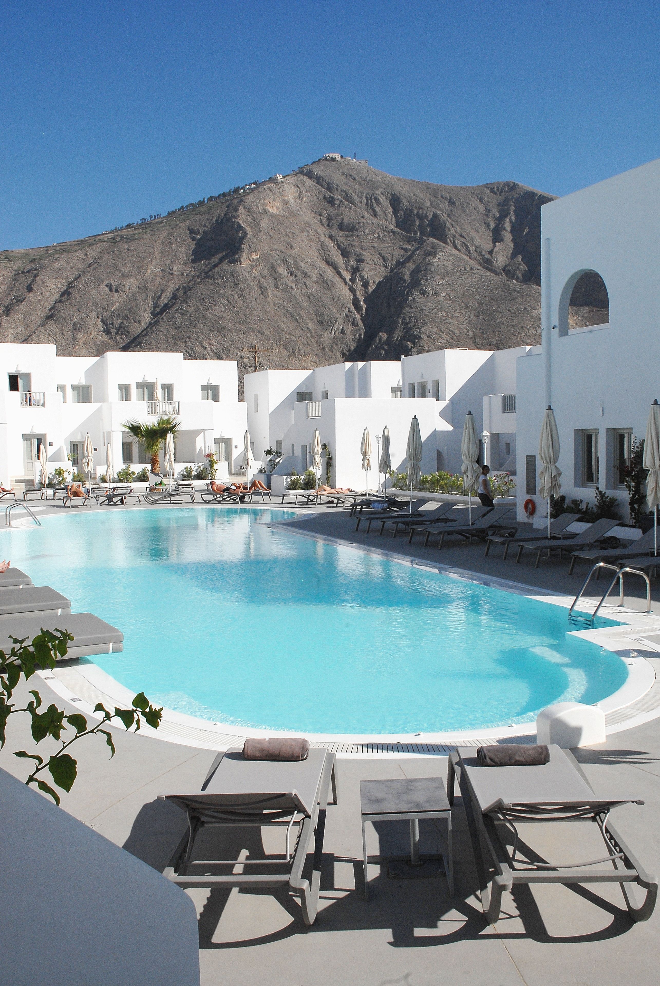 A pool area with lounge chairs and umbrellas, surrounded by white buildings with a mountain in the background.