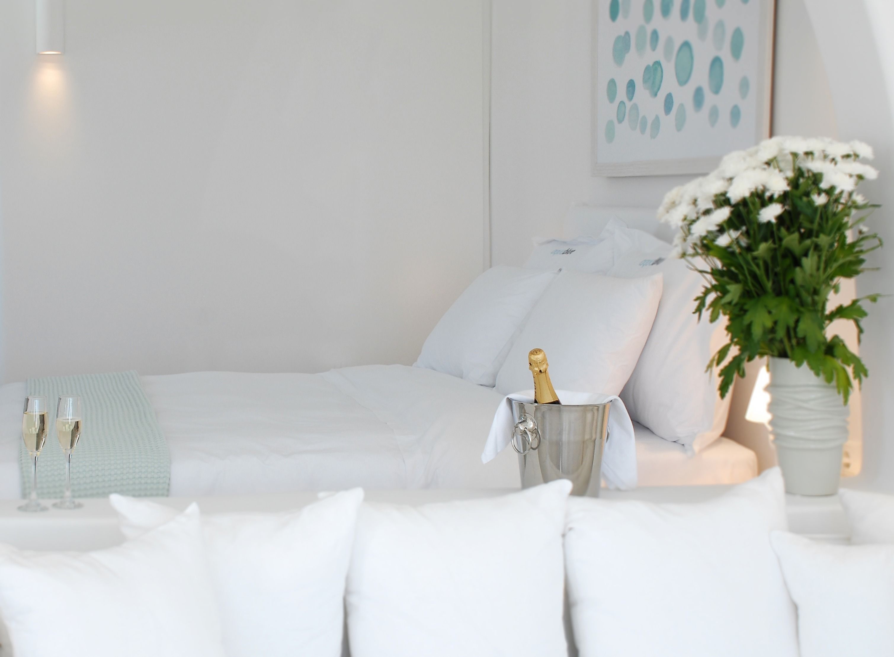 Un dormitorio ordenado con cama, almohadas, una mesita de noche con una botella de champán y un jarrón con flores.