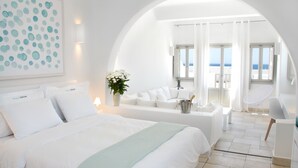 A modern hotel room with a large bed, a sitting area with a view of the sea, and a balcony with white curtains. - Aqua Blue Hotel (Santorini)