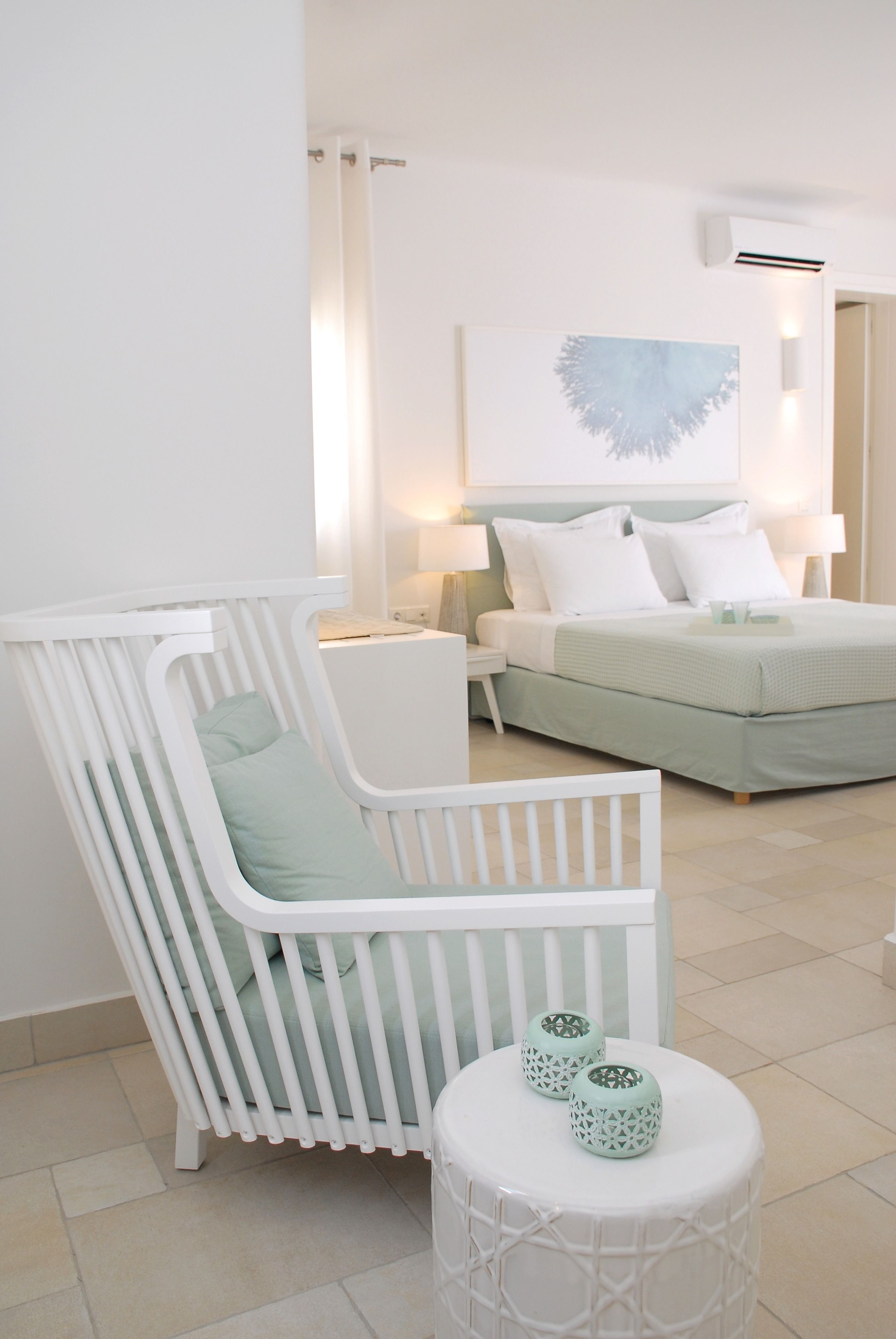A baby crib with a light green cushion, a white bedside table with a lamp, a bed with white bedding, and a wall-mounted air conditioner.