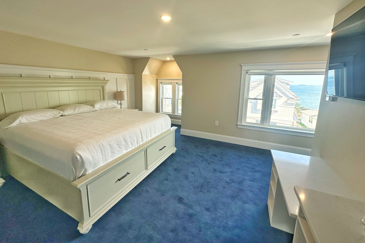 A bedroom with a large bed, a desk, a window with a view of the sea, and a ceiling with recessed lighting.