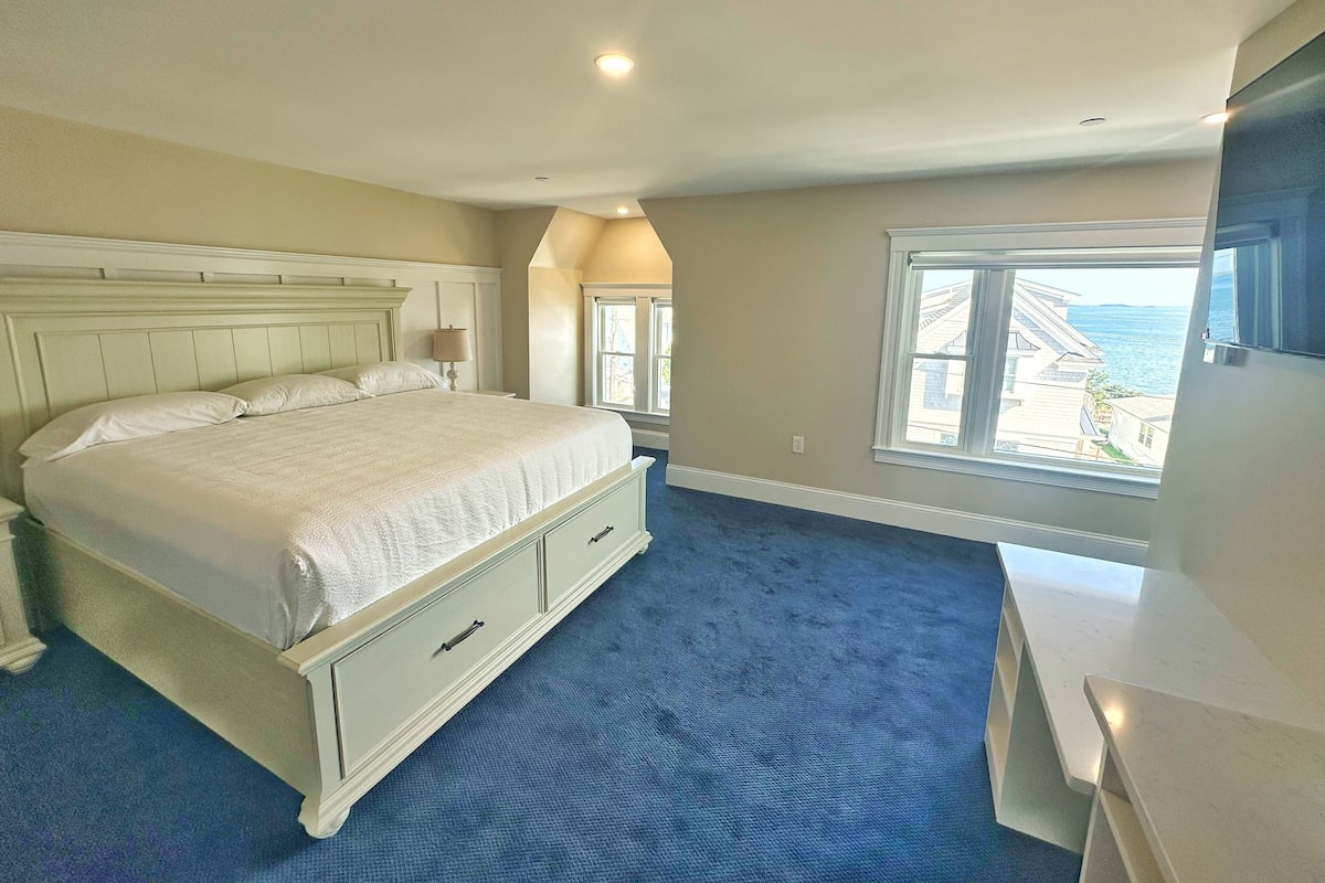 A bedroom with a large bed, a dresser, a nightstand, and a window with a view of the sea.